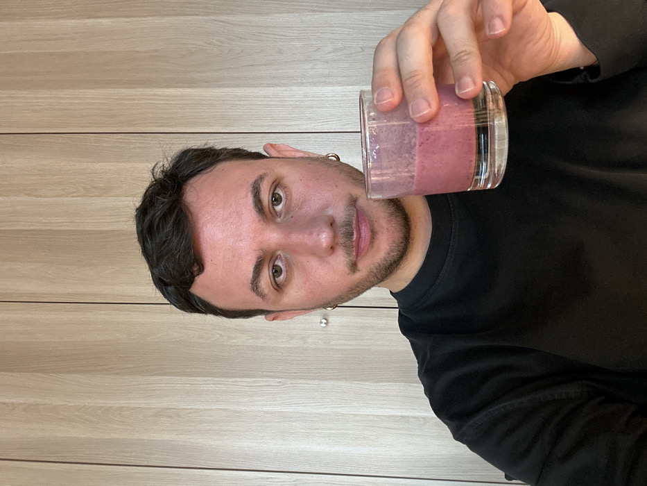 The image displays an adult male in a medium close-up, looking directly at the camera with a neutral expression. He has dark hair, a light complexion, and light facial stubble, wearing a black long-sleeved shirt. A gold hoop earring is visible in his right ear. In his left hand, he holds a clear glass containing a pinkish-purple liquid, consistent with a smoothie or beverage. The background consists of uniform light-toned wooden panels with vertical grain, indicating an indoor setting, likely a room with cabinetry or a paneled wall. There are no identifiable landmarks or distinguishing features to determine a specific location within London, United Kingdom.