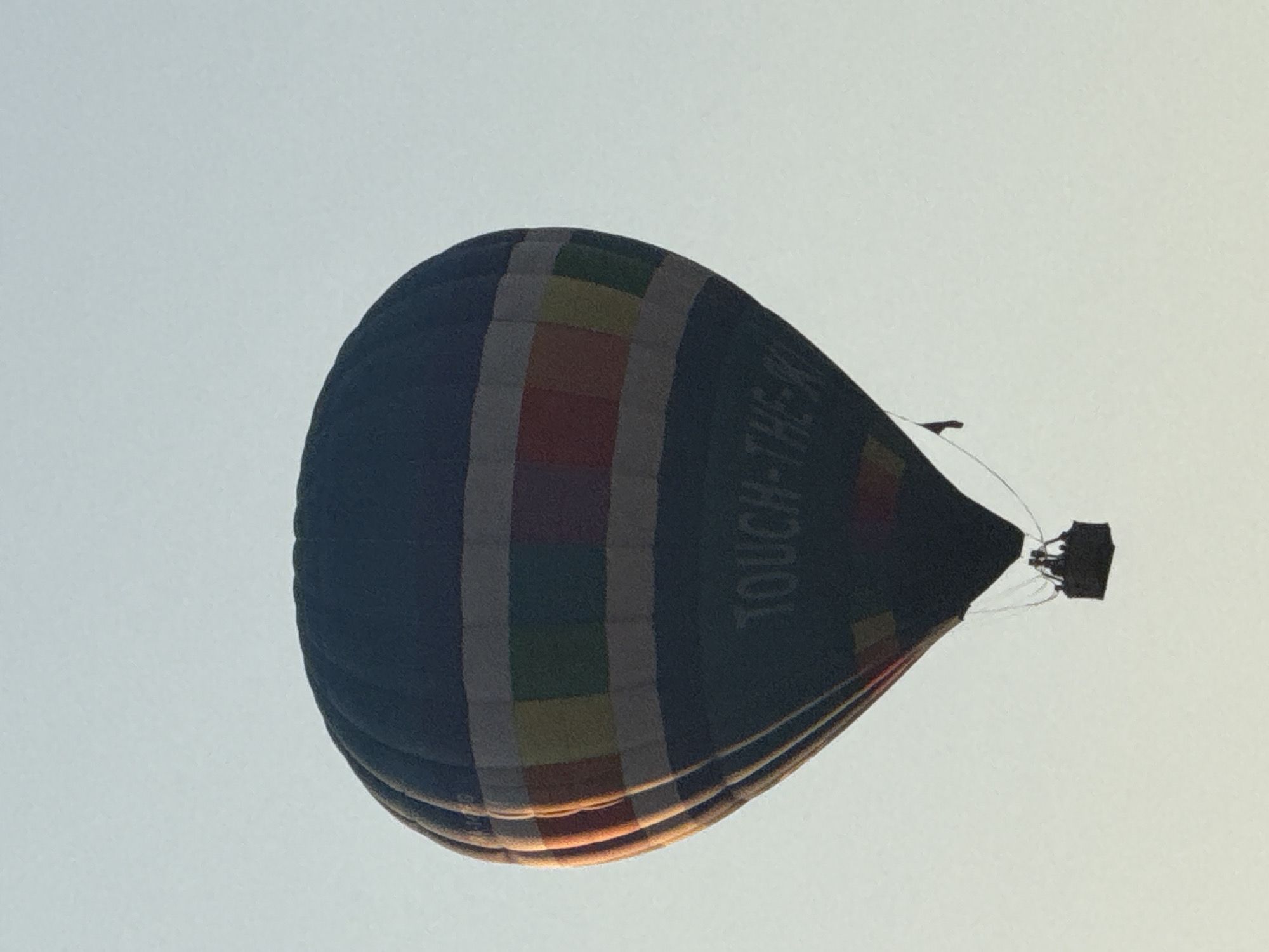 A hot air balloon with a dark blue envelope is depicted against a pale sky. The balloon features horizontal bands of colored rectangles: yellow, red, green, and grey. Text reading "TOUCH-THE-SKY" is visible in a lighter color on the lower portion of the envelope. A dark, rectangular basket hangs beneath the balloon, suspended by wires. The basket appears to contain at least three silhouetted figures. The image captures the balloon in mid-flight, suggesting an outdoor recreational activity. The exact location in Dmitrov, Russia, is not discernible from the image itself, as there are no identifying landmarks or geographical features present. The lighting suggests it is either early morning or late afternoon.