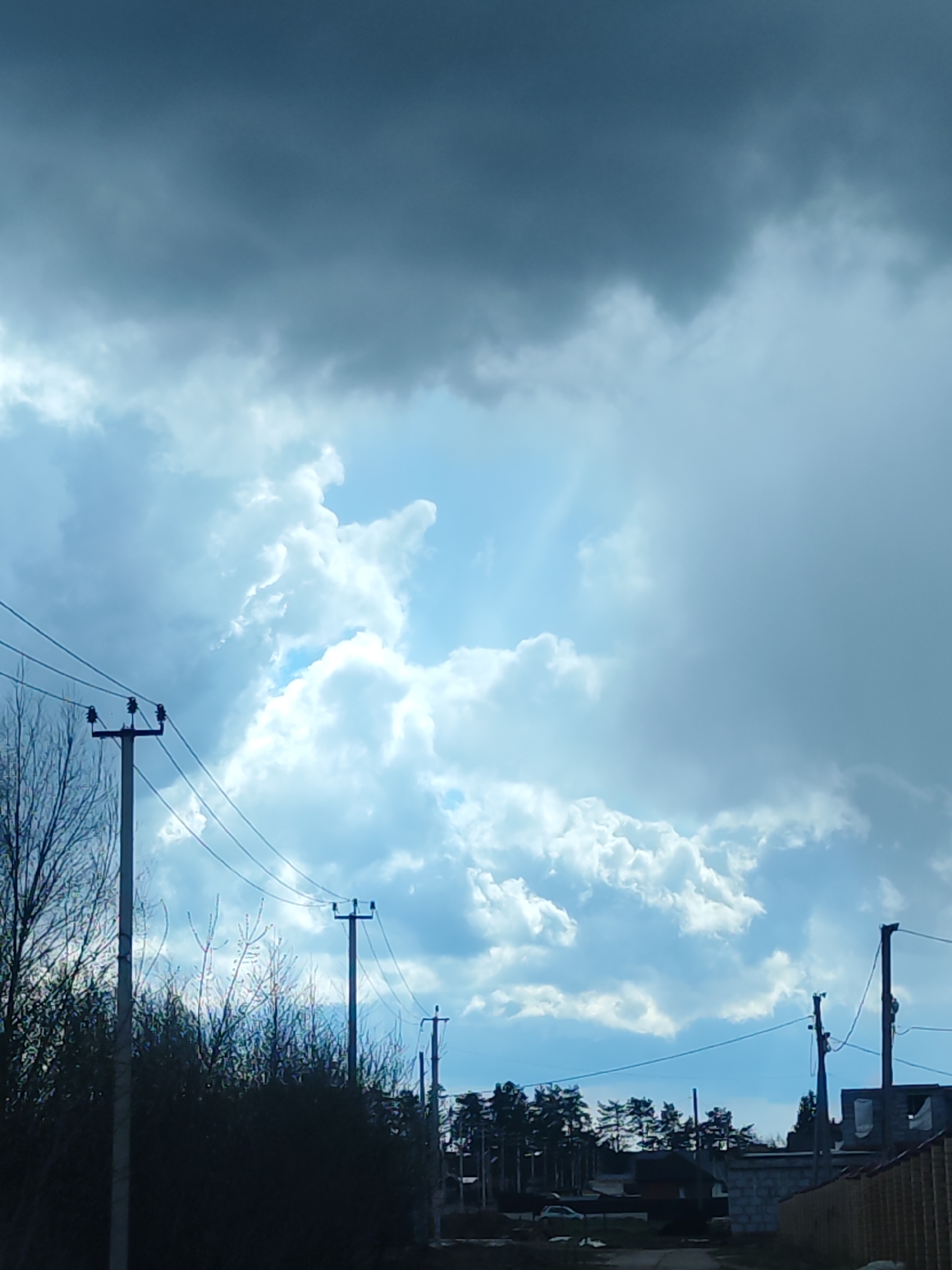 The image presents an outdoor scene in Chaikovsky, Russia, dominated by a dynamic sky. Heavy, dark gray clouds cover the upper part of the frame, while the mid-section and lower left reveal bright patches of blue sky and fluffy white cumulus clouds, suggesting sunlight breaking through. Rays of light are visible where the sun penetrates the cloud cover.
In the foreground on the left, there are bare branches of trees and dense, dark bushes. Extending across the midground are several utility poles with multiple overhead power lines. These poles are visible throughout the lower half of the image, receding into the distance on the right.
The background features a line of conifer trees, likely pines, beneath which are various residential structures. These include a building apparently under construction, indicated by visible concrete blocks, and a wooden fence. A dirt road or path is discernible in the lower center, with a light-colored car parked further down the road.
The scene depicts daytime with changeable weather, characterized by a mix of overcast conditions and clear, sunlit areas, possibly after or before precipitation. No discernible text is visible in the image.