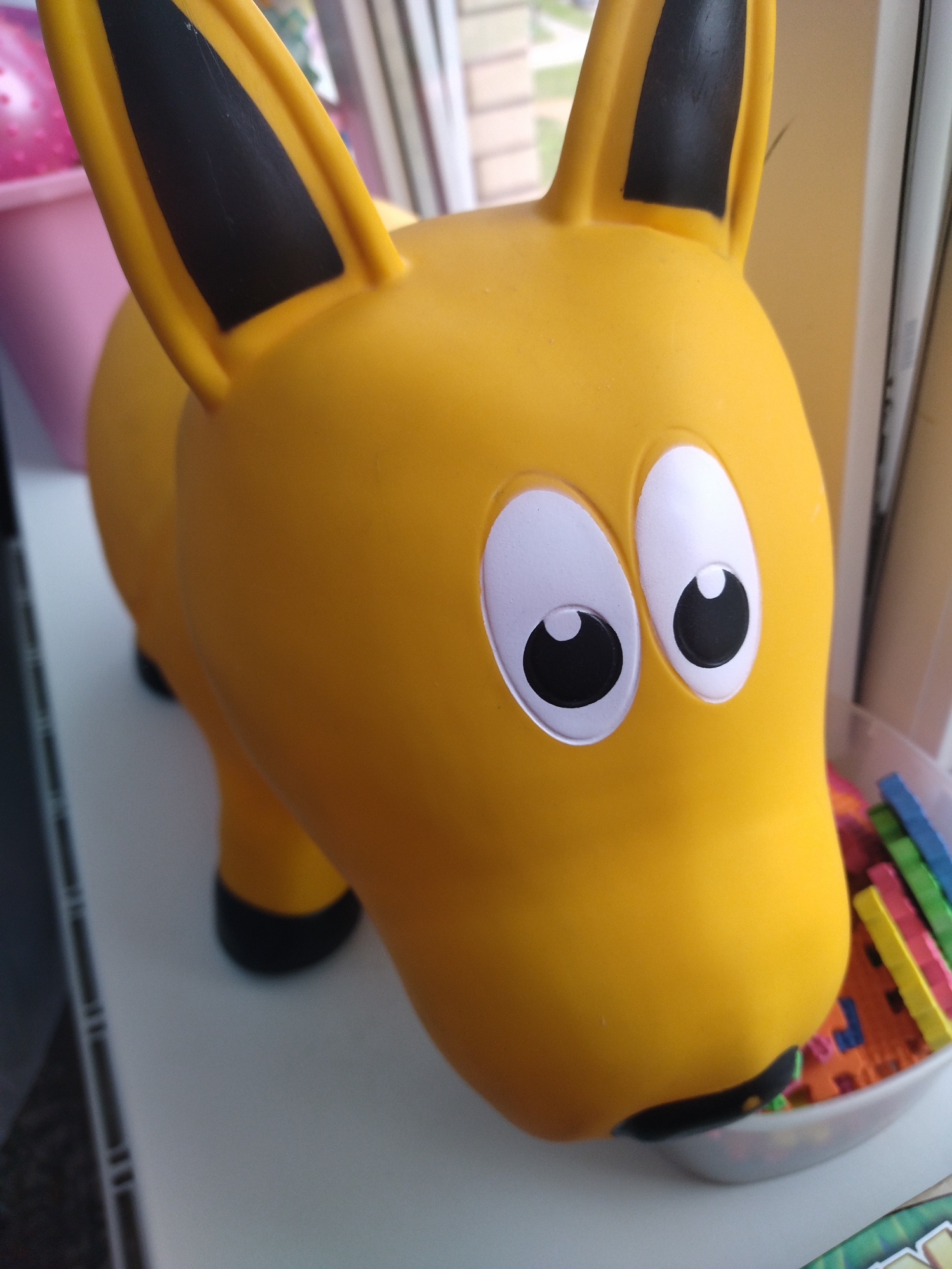 A close-up, eye-level, slightly tilted, bright outdoor shot captures a yellow rubber horse toy with large white eyes and black pupils, and black-tipped ears. The toy is positioned on a white surface, likely a table or shelf, with a blurred background that includes a pink bucket and window blinds. In the lower right corner, a clear plastic bowl contains a collection of colorful foam puzzle pieces. The scene appears to be an indoor environment. There are no people or animals present. The lighting is natural, suggesting daylight. The overall impression is one of a child's play area.