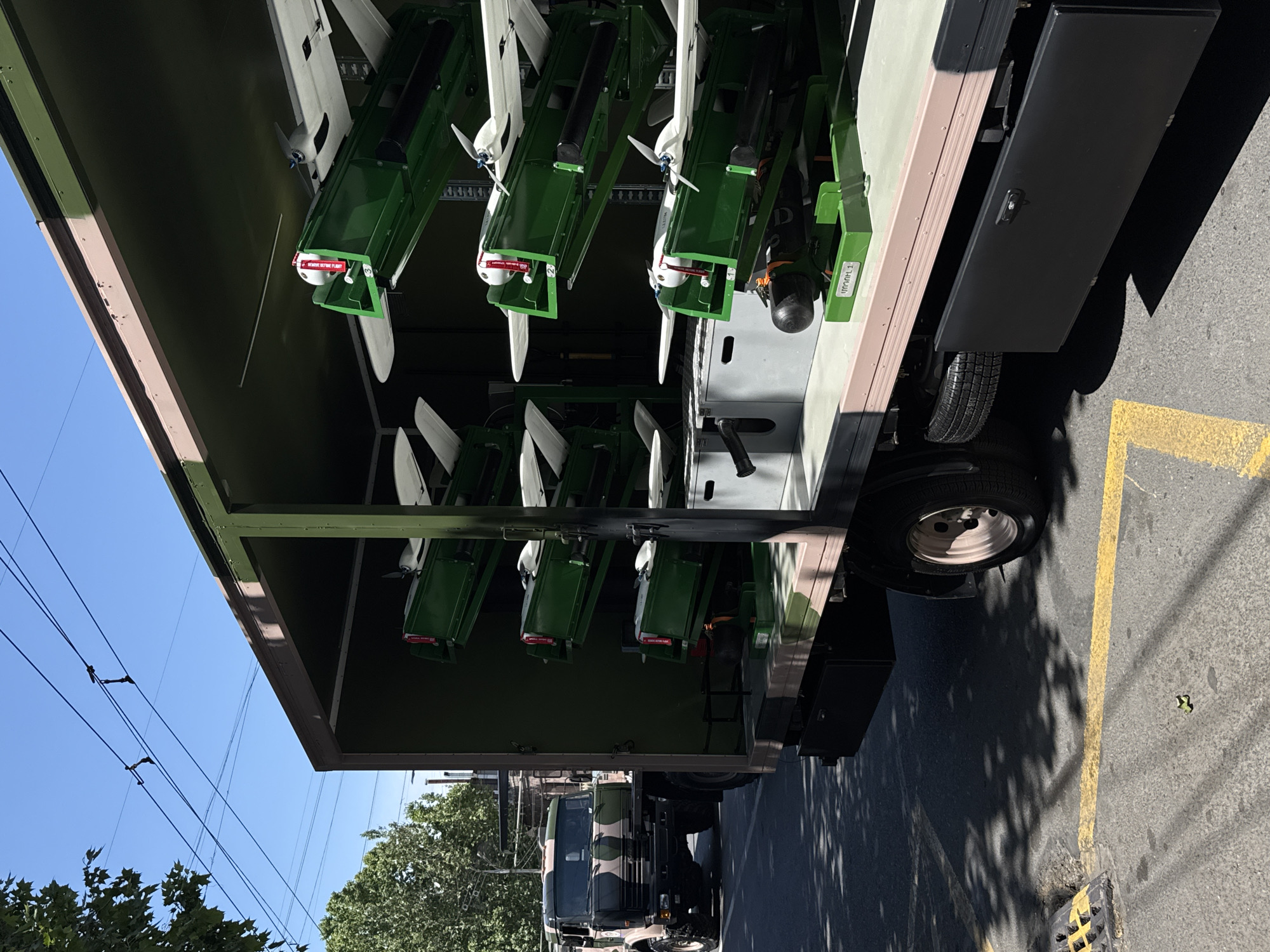 The image shows a military truck parked on a street, likely in Yerevan, Armenia. Inside the truck's cargo area, there are several green launch platforms, each holding what appears to be a small drone or unmanned aerial vehicle (UAV). The UAVs are equipped with propellers and have "REMOVE BEFORE FLIGHT" tags attached to their nose. The truck has a camouflage paint scheme. Power lines are visible in the upper-left of the frame. A second military truck can be seen further down the road. Yellow painted stripes are visible on the pavement.