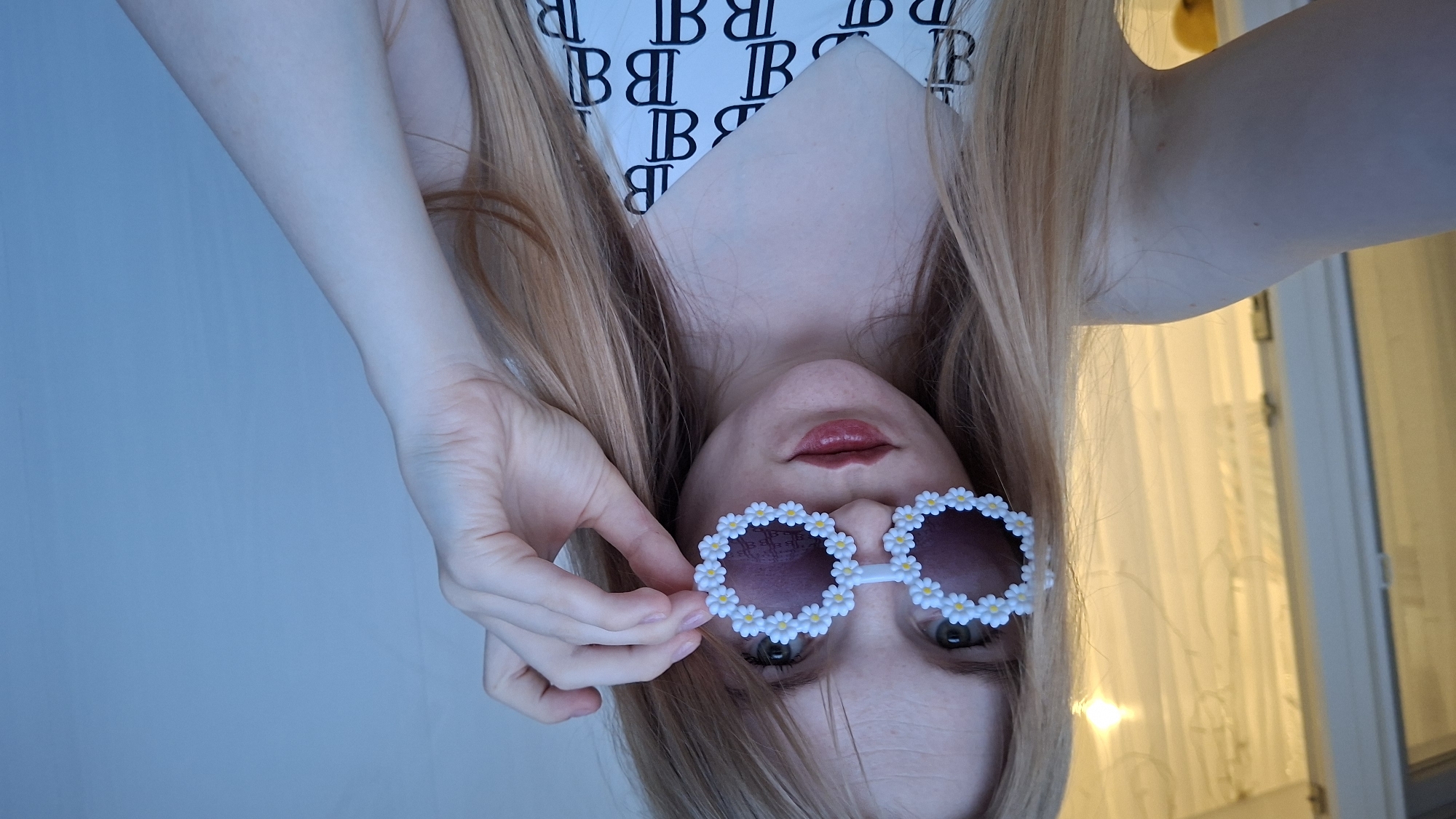 A young woman with long blonde hair is taking a selfie. She is wearing unique sunglasses decorated with small white flowers and has red lipstick on. She is wearing a white top with a repeating black "B" logo pattern. Her right hand is raised, adjusting the sunglasses. The background shows a sheer curtain and a wall. The image is a close-up shot and the location is not discernible from the visual information provided.