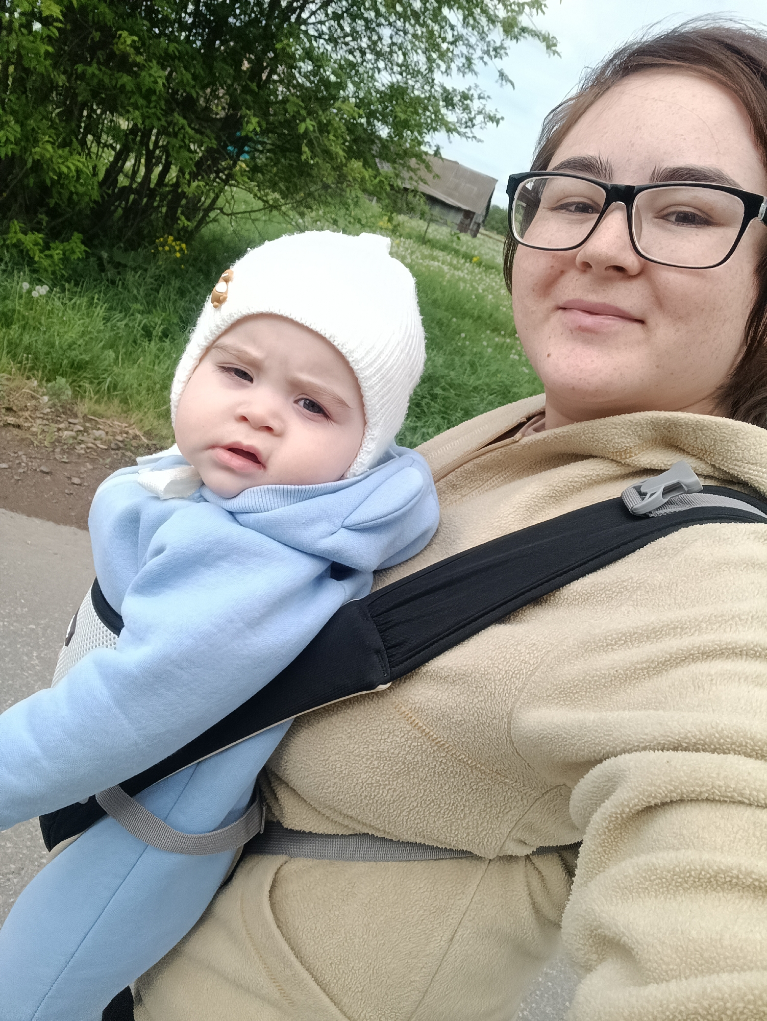 A young woman wearing glasses and a beige fleece jacket is standing on a paved path, holding a baby in a blue jumpsuit and white hat in a carrier. The baby is looking forward with a slightly stern expression. Behind them, there is green grass, trees, and a building in the background. The location is Jelpachiha, Russia, as indicated by the metadata of the image.