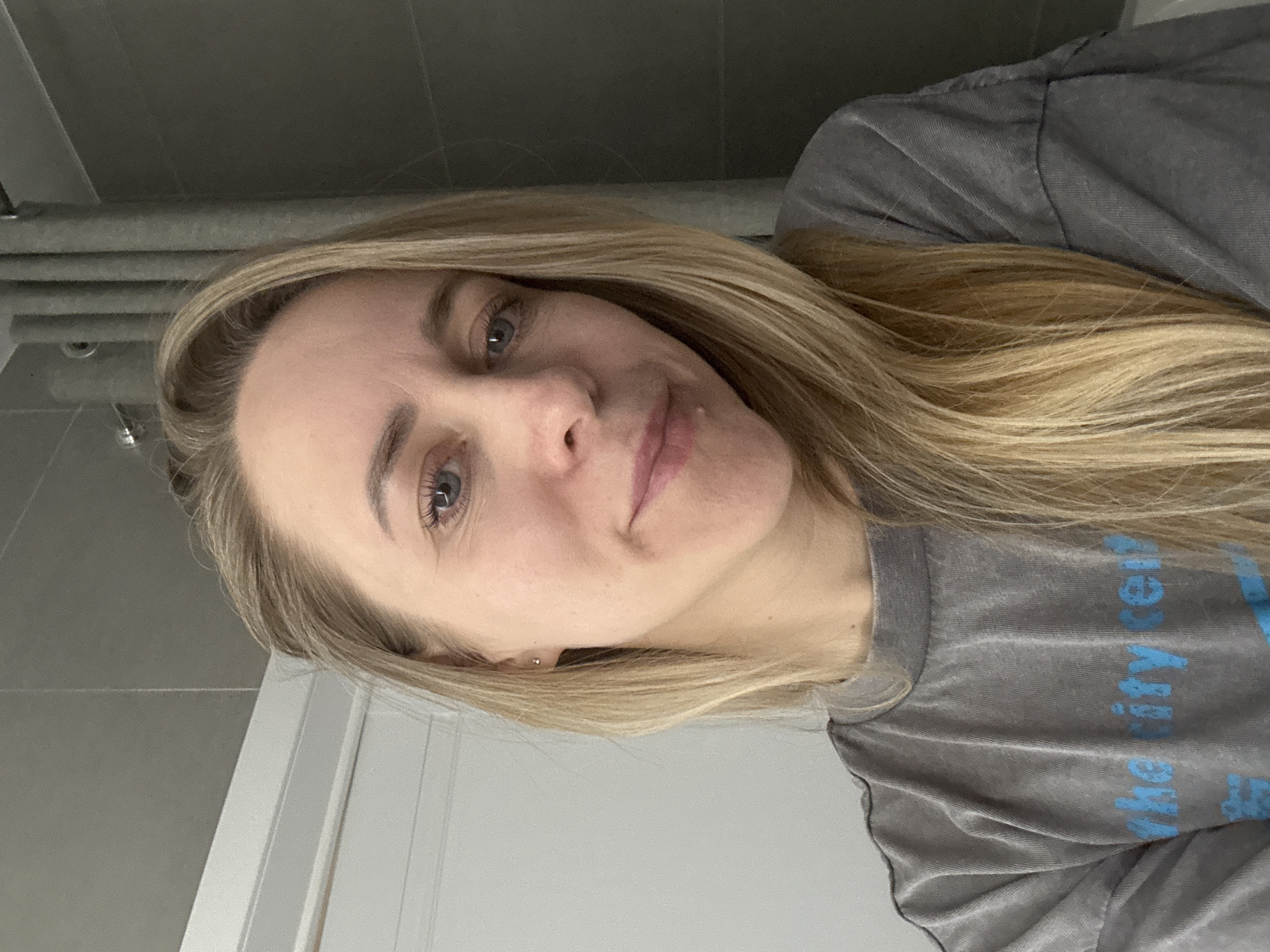 A self-portrait captures a young woman with long, straight, light blonde hair cascading over her shoulders. She has fair skin, blue eyes, and a neutral, gentle expression with a slight smile. She wears a relaxed, grey t-shirt that appears to be made of a soft, slightly textured fabric. Partial blue text is visible on the shirt, which, when upright, appears to spell out "the city re" or similar, with "rity" clearly visible in one of the crops. A small, shiny stud earring is visible in her right earlobe.
The image is taken indoors, with the woman looking directly at the camera, suggesting a selfie. The background to her left (top in the original image) consists of a muted grey wall or ceiling with visible pipes or structural lines. To her right (bottom in the original image), there's a section of a light-colored, possibly white, door or wall paneling. The lighting is soft and even, indicating an indoor setting during the daytime. The overall scene is simple and personal, focusing on the woman. The image is rotated 90 degrees counter-clockwise.