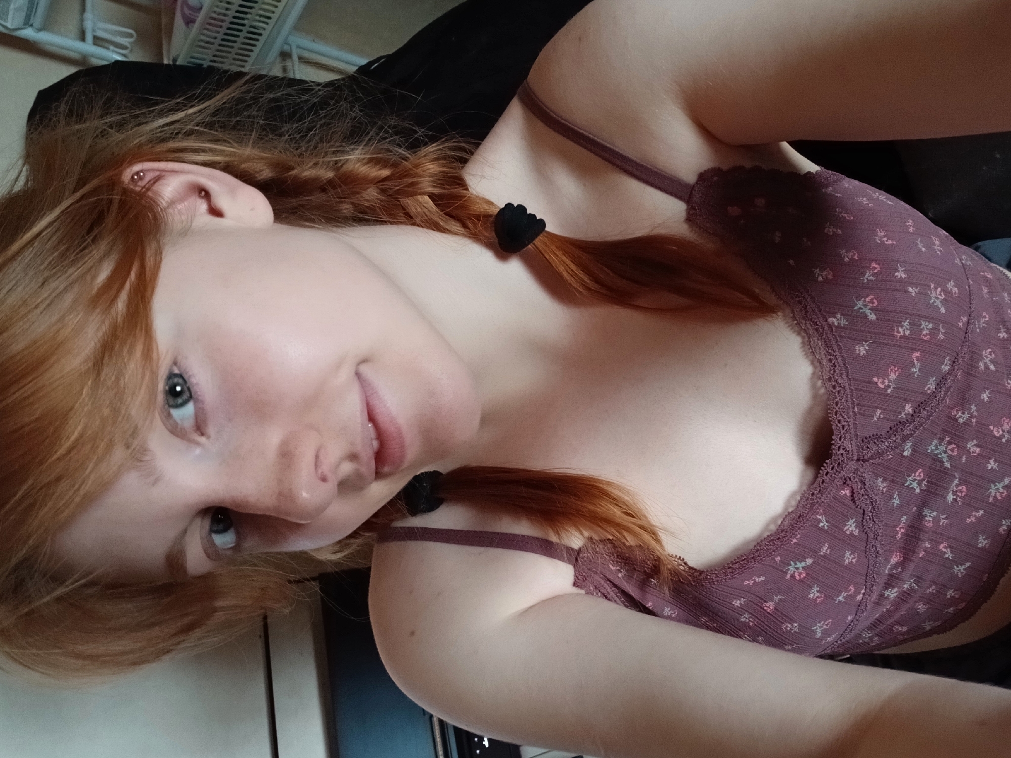 A young individual, appearing to be in Yekaterinburg, Russia, is captured in a close-up, upward-angled self-portrait. They possess fair skin, striking blue-green eyes, and long, reddish-brown hair styled into two braids. Each braid is secured with a small, black, hand-shaped hair tie. Visible on one ear are two piercings: a stud in the lobe and a barbell in the upper cartilage.
The person is wearing a deep purple top, possibly a bralette or camisole, featuring a delicate floral pattern in pink and lighter shades. The garment has intricate lace trim along its edges and thin spaghetti straps visible on the shoulders. The individual's expression is one of gentle contentment, with a soft, closed-mouth smile directed towards the camera.
The setting appears to be indoors, with soft, diffused lighting. The background is largely out of focus, showing dark fabric, possibly a pillow or clothing, and a hint of a lighter surface or wall, suggesting a relaxed, casual environment such as a bedroom or living space. There is no visible text within the image.
