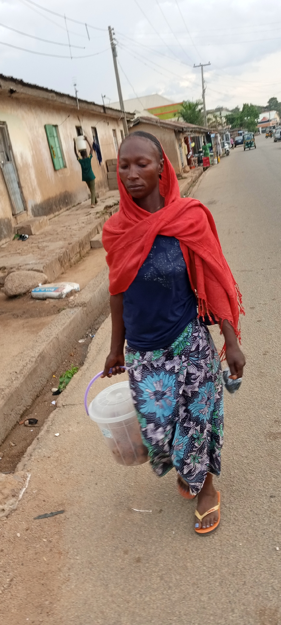 In Jos, Nigeria, a woman walks along a paved road. She wears a red scarf draped over a dark blue top and a floral print skirt. She is carrying a translucent bucket with a purple handle in her left hand. She wears orange flip-flops. In the background, another person carries a container on their head. Buildings and a road with vehicles are visible in the distance.