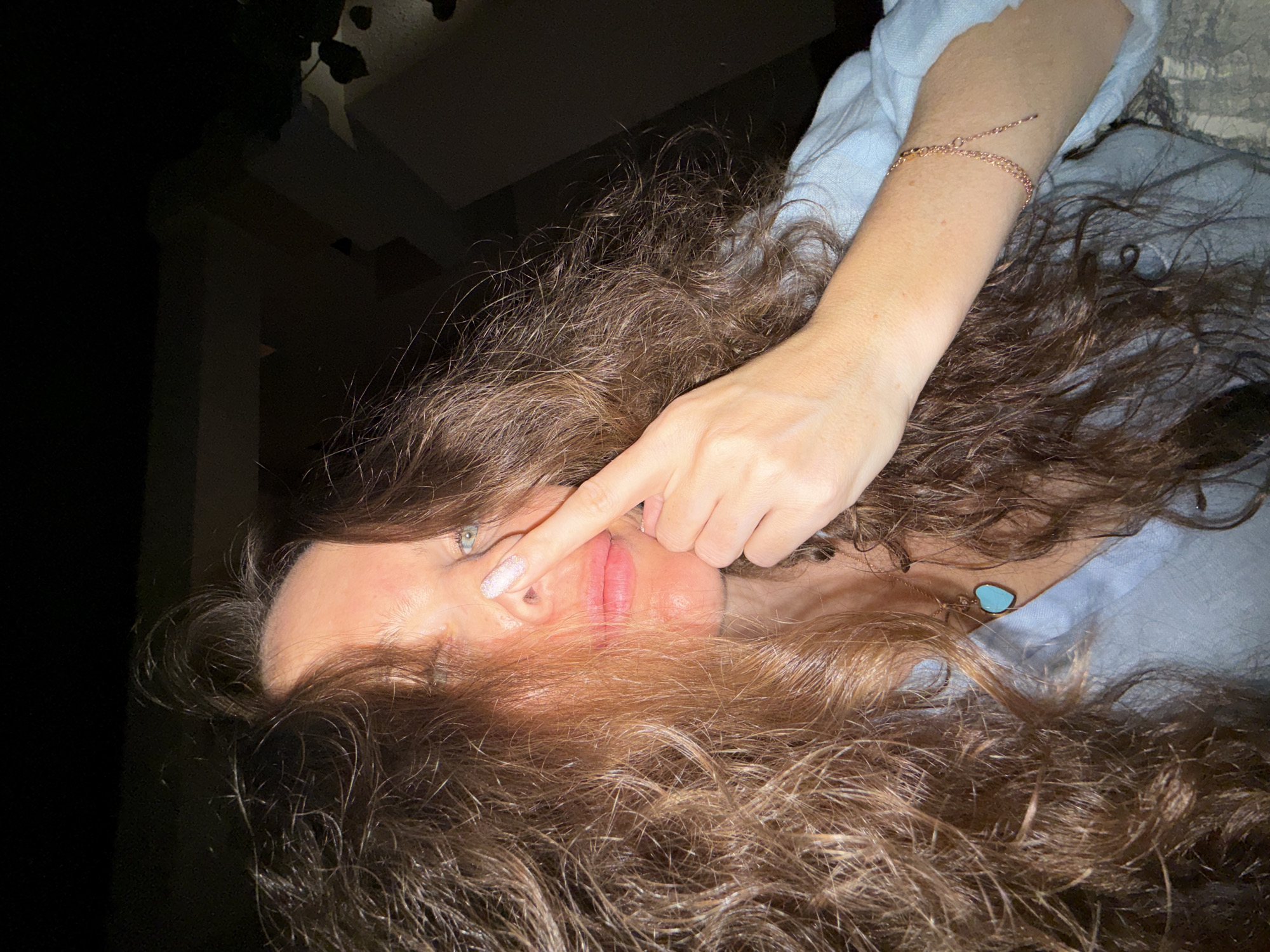 The image shows a close-up of a woman's face, with her right index finger touching her nose. She has long, curly brown hair that partially obscures her face. She is wearing a light blue shirt and a necklace with a blue heart-shaped pendant. A thin gold bracelet is visible on her right wrist. The background is dark and out of focus, suggesting an indoor or nighttime setting. There are no discernible landmarks or features that would indicate a specific location within Dubai, United Arab Emirates.