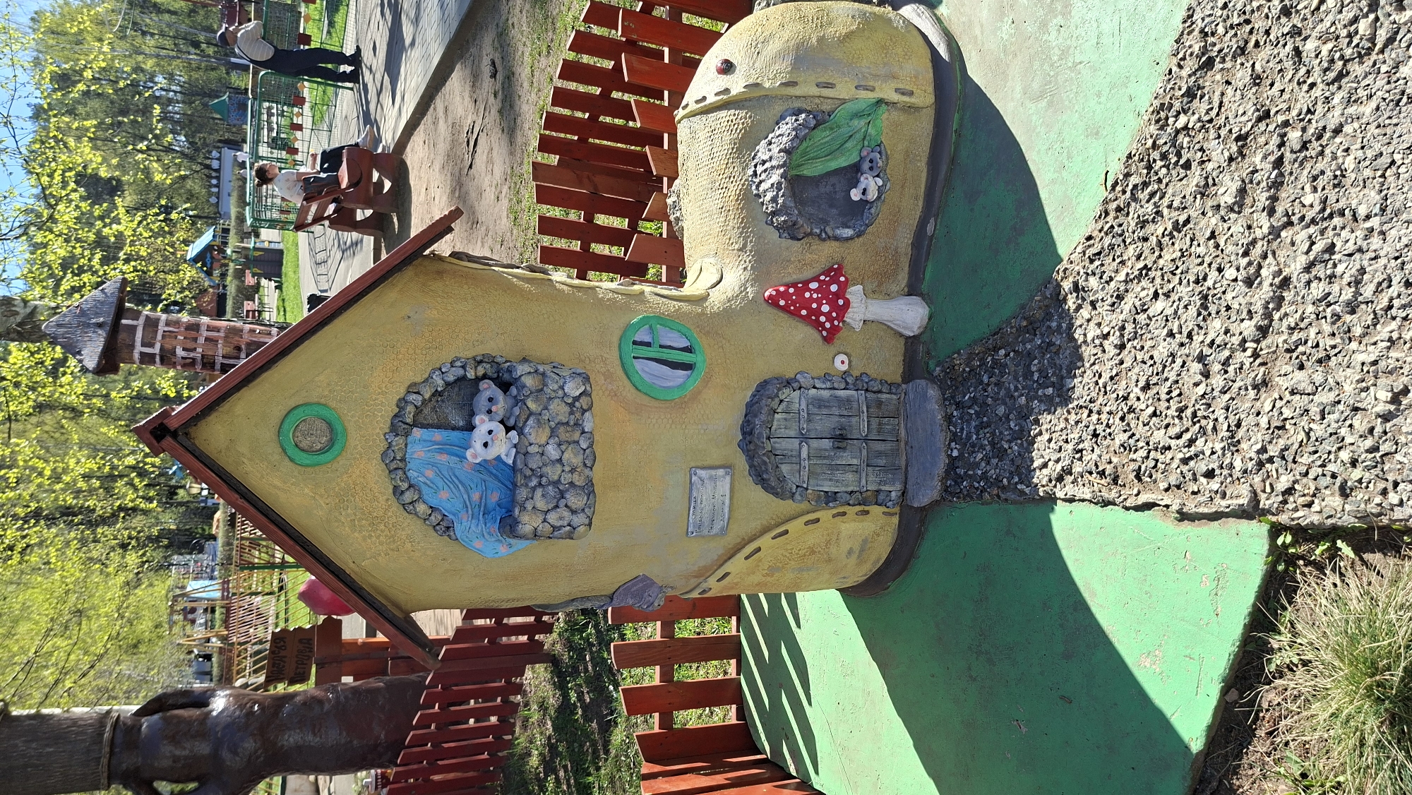 The image shows an outdoor scene at the Children's Park in Chaikovsky, Russia. The main subject is a whimsical, boot-shaped playhouse painted in yellow hues. It features faux windows with representations of small animals, a door, and a mushroom embellishment. The playhouse is situated on a green painted surface.
In the background, people are visible on benches and walking in the park. Trees with fresh green foliage surround the area. There is a red wooden fence nearby and a tall cylindrical structure resembling a chimney.