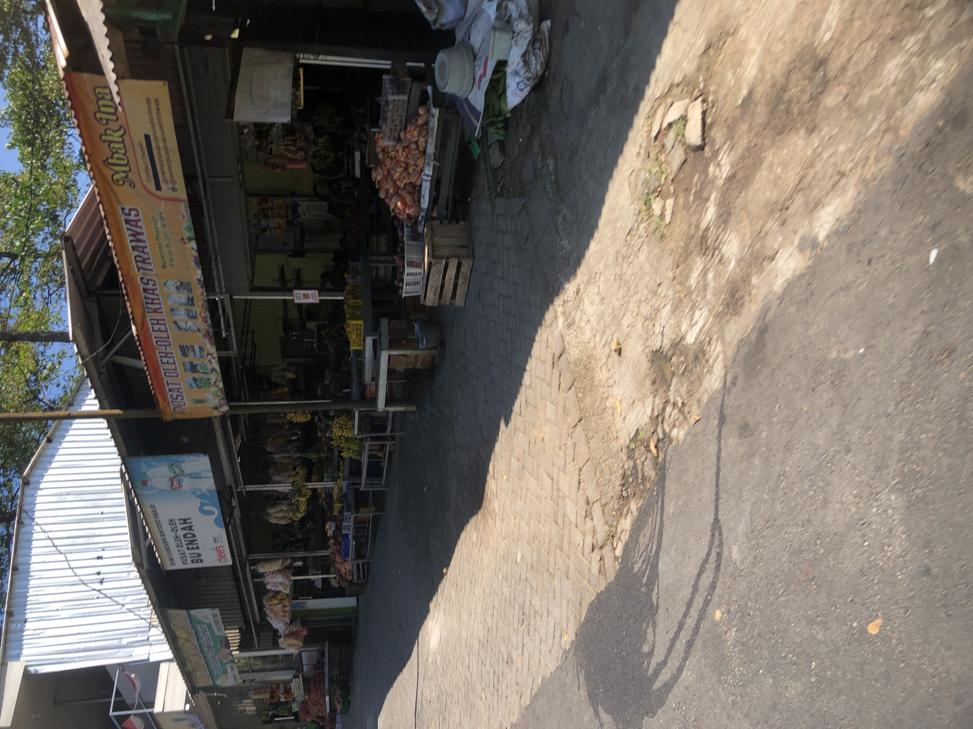 The image depicts a street scene in Trawas, Indonesia, likely a marketplace or a row of shops. A building with a white corrugated metal roof is visible on the left. Several stalls are set up along the street, displaying a variety of goods, including fruits like bananas and what appear to be onions or potatoes. Banners are hung above the stalls, with text in Indonesian. One banner reads "Pusat Oleh-Oleh Khas Trawas" which translates to "Trawas Souvenir Center," and another displays "Mbak Ina." The street itself is paved with interlocking stones and asphalt, with shadows cast by the sun indicating it is daytime. There are no people clearly visible in the image, but the presence of the stalls suggests commercial activity. The environment appears to be outdoors, with trees visible in the background.