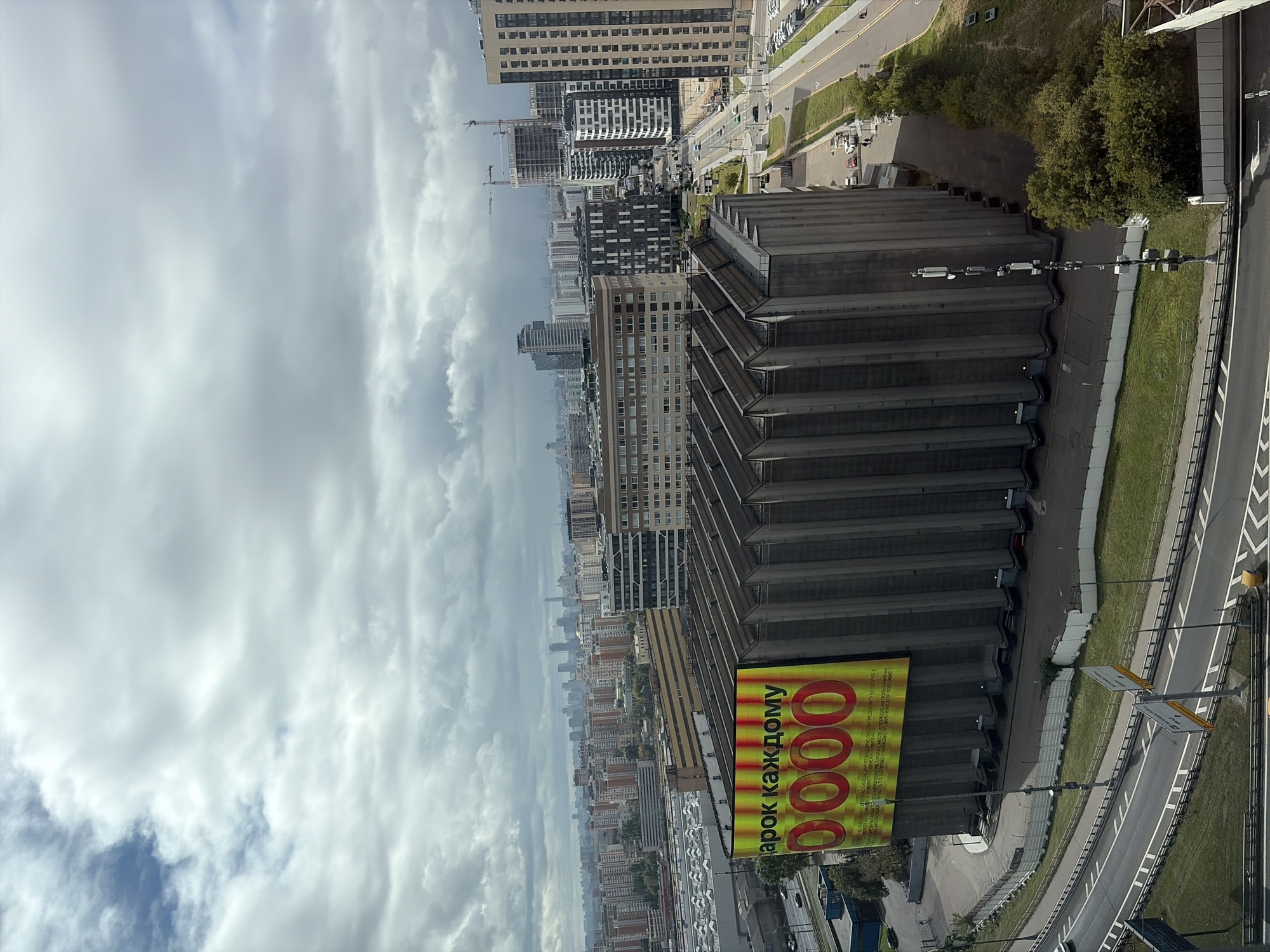 The image is an aerial view of an industrial or commercial area in Moscow, Russia, under a cloudy sky. In the foreground, a massive, ribbed concrete structure, possibly a grain silo or industrial building, dominates the scene. A large, bright yellow billboard is attached to its side, featuring large red numbers "0000" and Russian text that reads "Подарок каждому" (A gift for everyone). Surrounding this structure is a complex network of roads, some with traffic, and surrounding buildings of various sizes and architectural styles, including residential high-rises and modern office buildings. Construction cranes are visible in the distance, suggesting ongoing development in the city. Green spaces and trees are interspersed with the urban landscape.