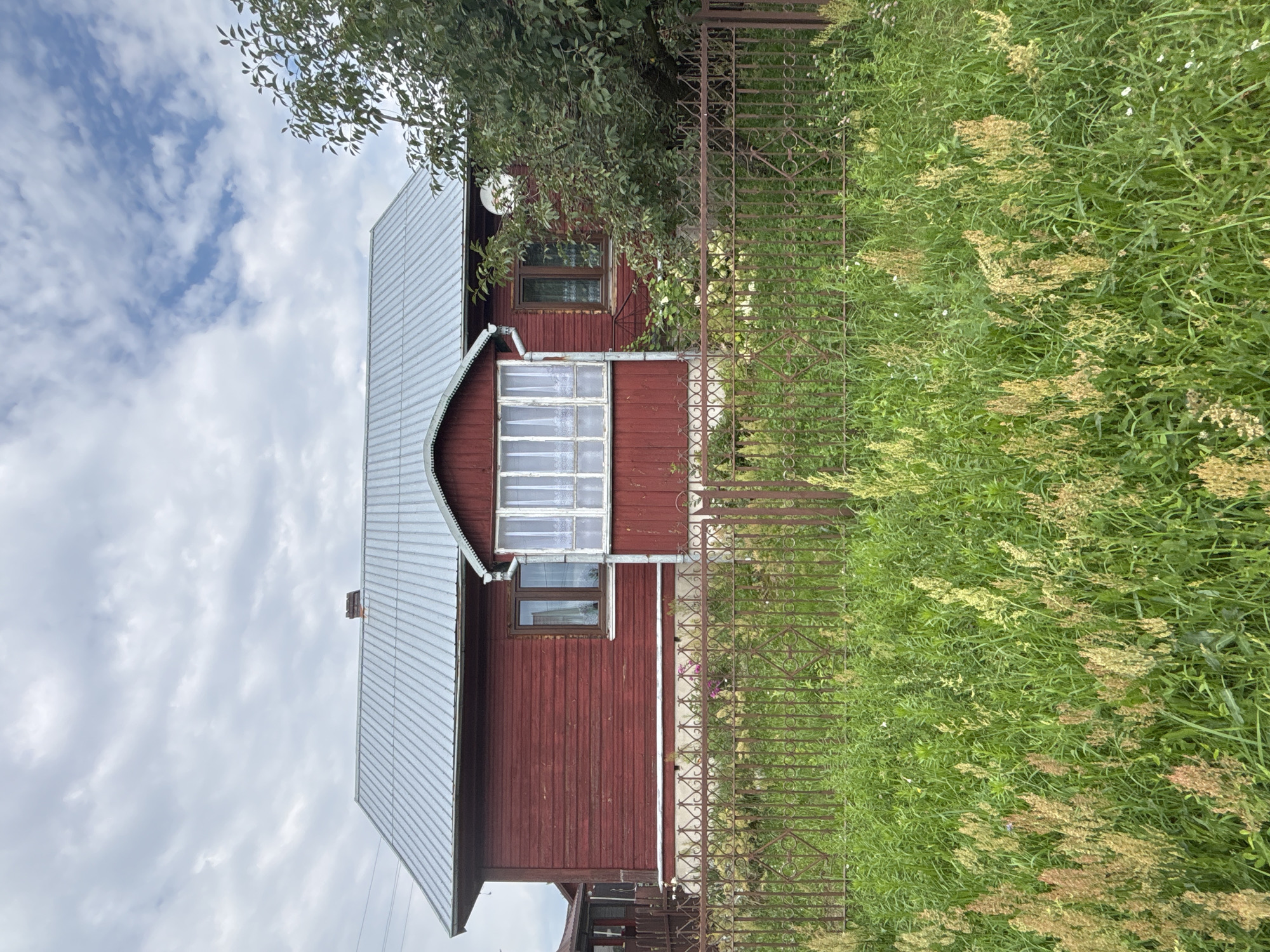 A red wooden house with a metal roof is set back from the road, surrounded by a low metal fence. The house has several windows, some with white curtains visible. A small porch with windows is located in the center of the house. Tall green grass and wildflowers grow in front of the fence. The sky above is partly cloudy, with patches of blue visible. The scene is situated in Trześcianka, Poland, a village known for its rural character and traditional wooden architecture. The overall impression is one of a modest, well-maintained rural dwelling.