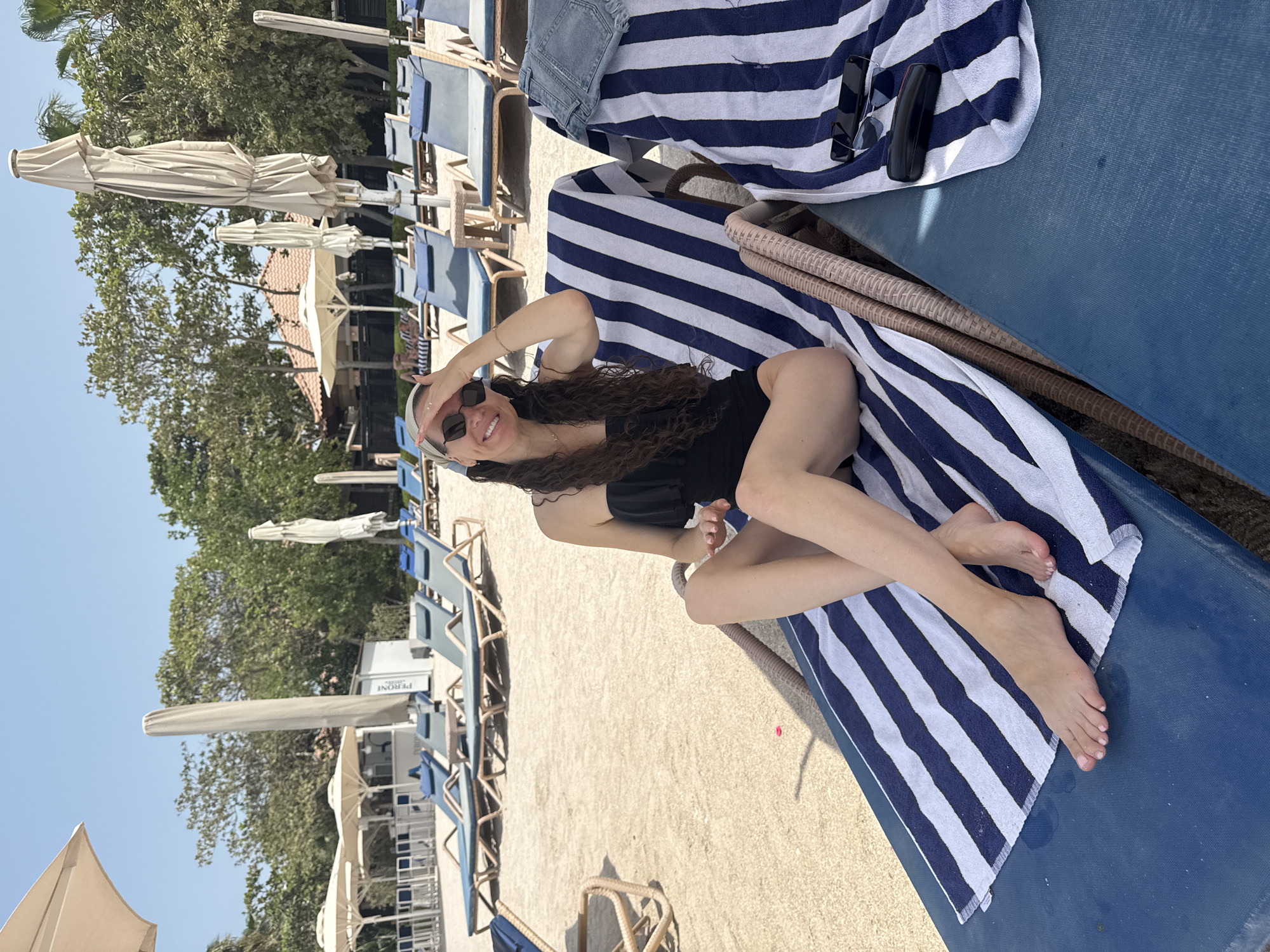 A woman is relaxing on a beach chair on a sandy beach, a common scene in Dubai, United Arab Emirates. The woman is wearing a black swimsuit and sunglasses, and she has her hand on her forehead, shielding her eyes from the sun. She is smiling and appears to be enjoying herself. In the background, there are several other beach chairs and umbrellas, as well as some palm trees and buildings. The sky is blue and clear, indicating good weather. The overall impression is one of a relaxed and enjoyable day at the beach in a popular tourist destination.