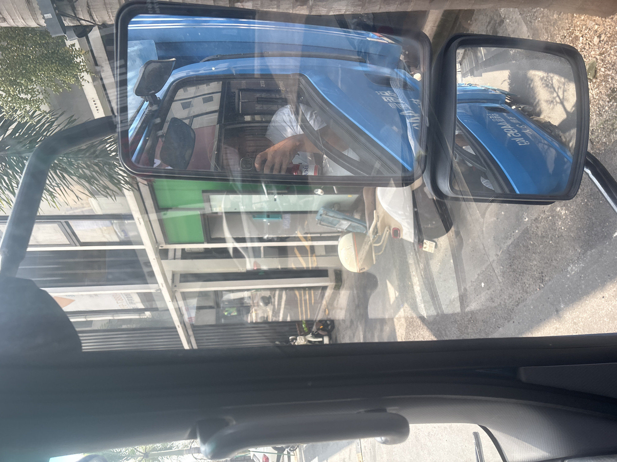 This image shows a view from inside a vehicle, looking out through the windshield and side mirrors. The scene appears to be in a city, possibly Baiyun District, China, given the presence of buildings with Asian architectural elements and signage that may be in Chinese.
In the main windshield view, we see a street with a sidewalk and a building facade. A white car is parked on the left, and a scooter is parked further down the street. A person is reflected in the side mirror on the right, holding a phone and taking a selfie. This person is wearing a white t-shirt. The vehicle's interior, including part of the dashboard and door, is visible in the foreground.
The side mirrors provide further reflections. The larger side mirror shows the driver's side of a blue truck or van, with text in what appears to be Chinese characters on its side. A smaller mirror above it reflects the side of the same truck. The background visible through the windshield and in the mirrors includes buildings, trees, and shadows cast by sunlight. The overall impression is that of a typical urban street scene in China, with traffic and pedestrians.