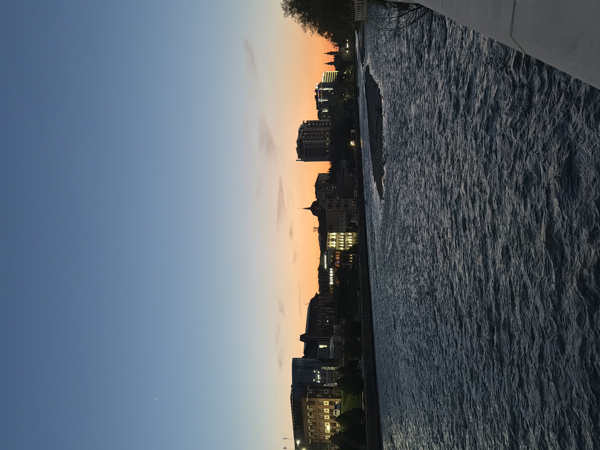 The image captures a cityscape at dusk along a river. Buildings are visible in silhouette against a gradient sky transitioning from blue to orange, with lights on in some windows. The river occupies a significant portion of the right side of the image, its surface reflecting the fading light. There is an indication of a structure on the right side that may be a bridge. The location is assumed to be Chermen, Russia based on the query. There are no people visible in the image.