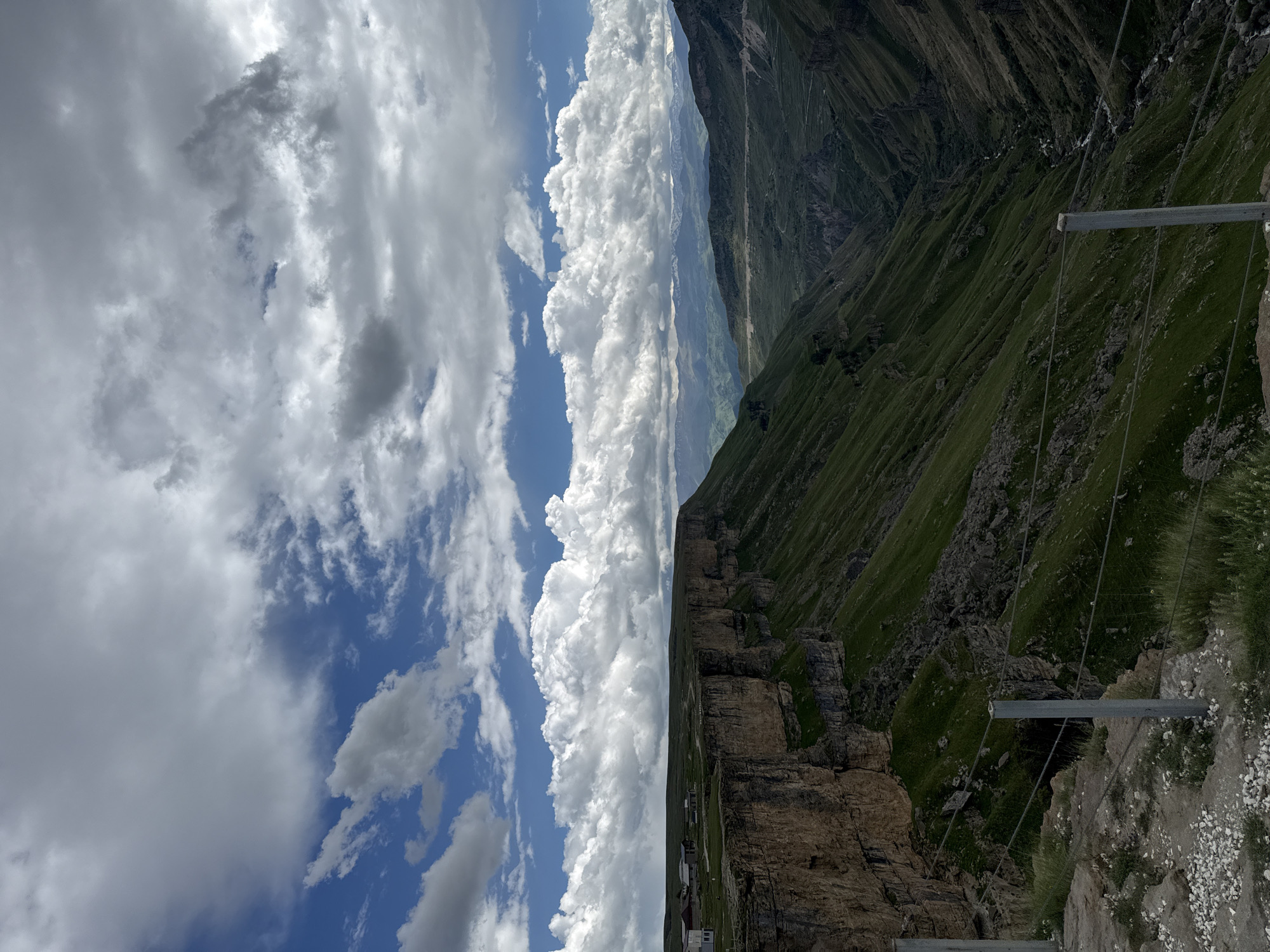 This panoramic image presents a dramatic mountainous landscape, likely situated in Khunzakh, Russia. The scene is dominated by a vast sky on the left and upper portions, featuring a mix of brilliant blue patches and large, white, fluffy cumulus clouds, with some darker, grey clouds hinting at a dynamic atmosphere.
To the right, a steep, rugged cliff face of brown and grey rock descends sharply. Below and extending into the distance are rolling green mountains and hills, thickly covered in verdant grass, interspersed with rocky outcrops. In the immediate foreground on the bottom right, a man-made structure is visible: a horizontal metal beam supported by several tension cables that stretch downwards and outwards, likely forming part of a safety barrier or a viewing platform. A gravelly path is also visible here.
In the mid-ground, at the base of the cliff, small buildings or a settlement can be seen, indicating human habitation amidst the imposing natural environment. Further in the background, layers of mountains fade into the horizon under the expansive sky.
The photograph was taken during daytime, likely late morning or early afternoon, given the bright illumination and distinct shadows. The weather appears to be partly cloudy with significant cloud formations. The image conveys a sense of grandeur, natural beauty, and the immense scale of the mountain terrain. No visible text is present.