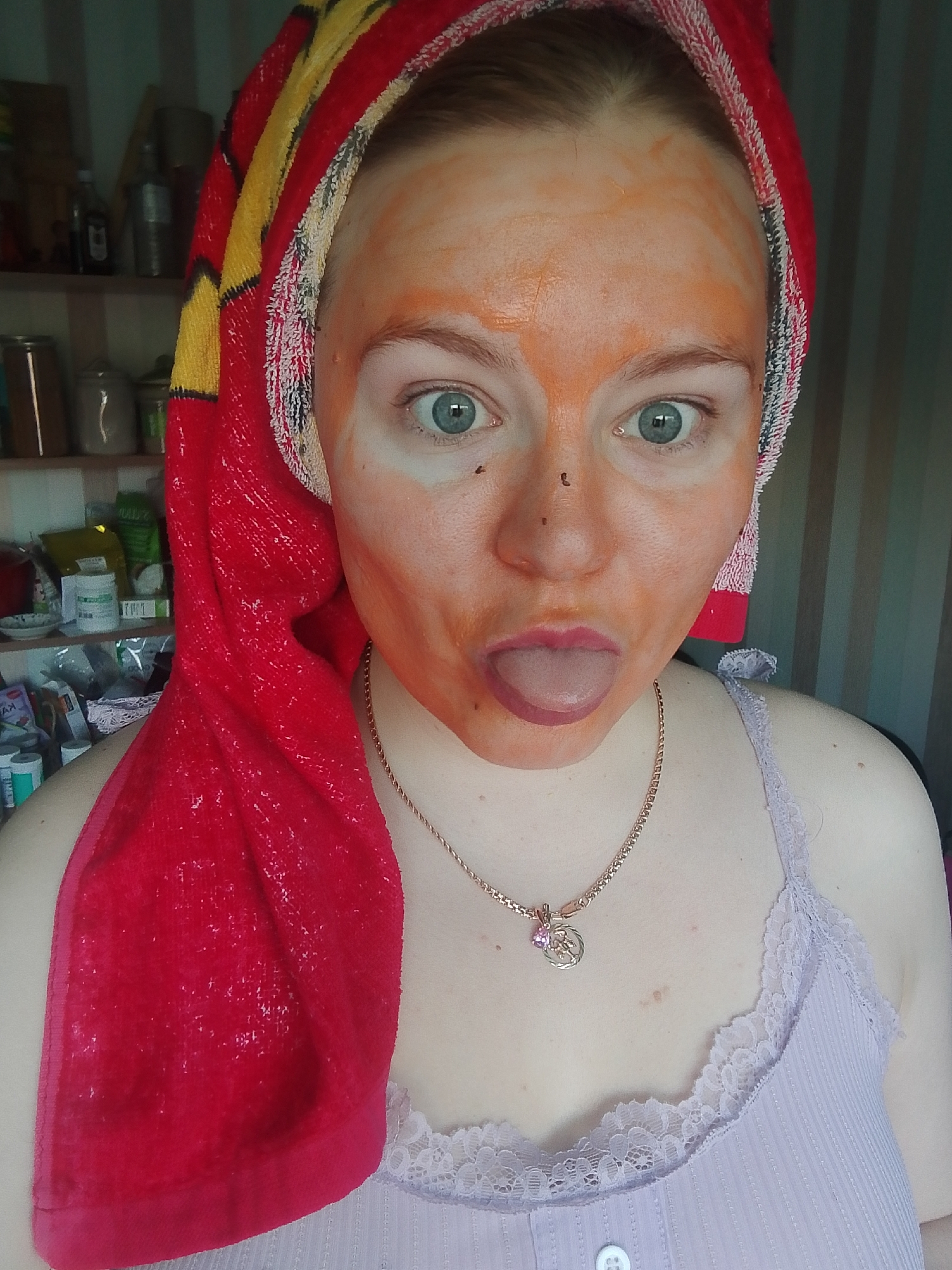 A woman is centered in the frame, wearing a lavender tank top with white lace trim and a gold chain necklace with a pendant. A red and yellow towel is wrapped around her head. Her face is covered in an orange mask, and she is sticking out her tongue.
The background shows a shelf cluttered with jars and containers, suggesting a kitchen or pantry. The scene appears to be indoors. The photo was not necessarily taken in Izhevsk, Russia, as there are no contextual clues.