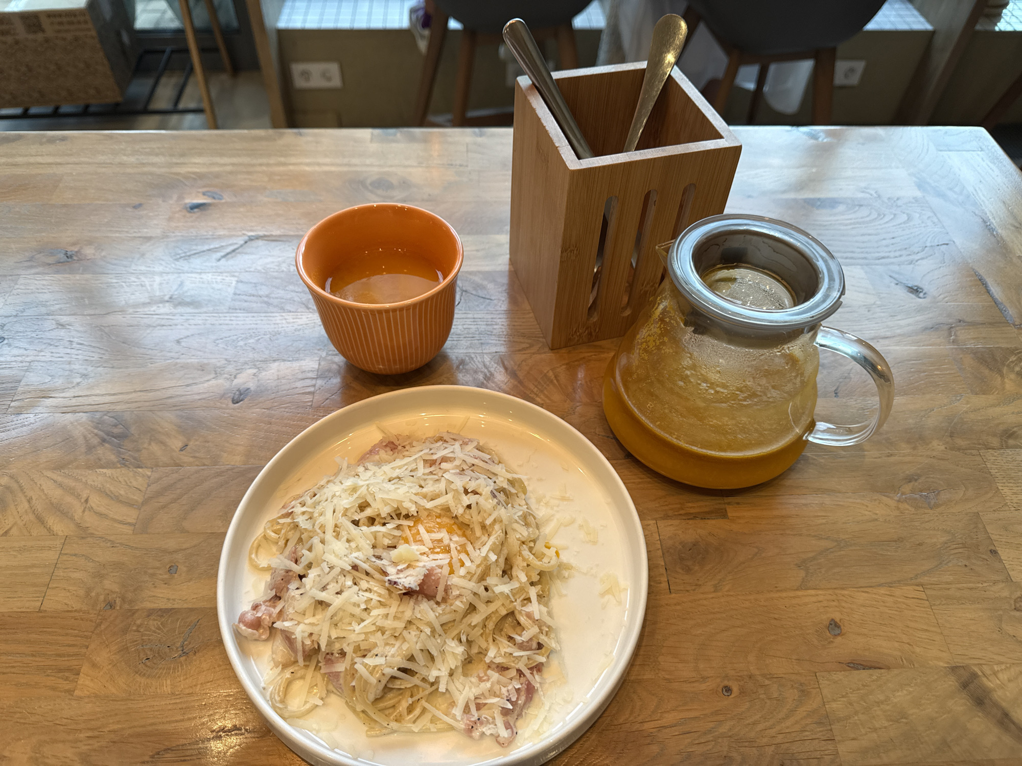 A plate of spaghetti carbonara is placed on a wooden table, topped with shredded cheese and a partially visible egg yolk. Next to the plate, an orange ceramic mug holds a clear, amber liquid. To the right, a glass teapot contains the same amber liquid, topped with a silver lid. A wooden utensil holder sits behind the mug and teapot, containing two silver utensils. The scene is set in a cafe or restaurant, with blurred chairs and tables visible in the background. The lighting suggests it could be daytime, possibly late morning or early afternoon. The overall mood is casual and relaxed, with the focus on the meal. No visible text is present in the image.