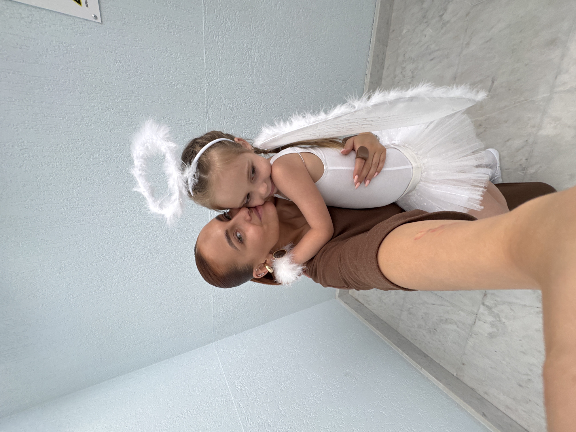 A woman in a brown shirt and gold hoop earrings is lying on her back on what appears to be a massage table in a room with light blue walls and a white marble floor. A young girl, dressed as an angel with white wings, a halo, and a white tutu, is lying on top of her, resting her head near the woman's face. The photo is taken with a selfie angle. The location is possibly Chaikovsky, Russia.