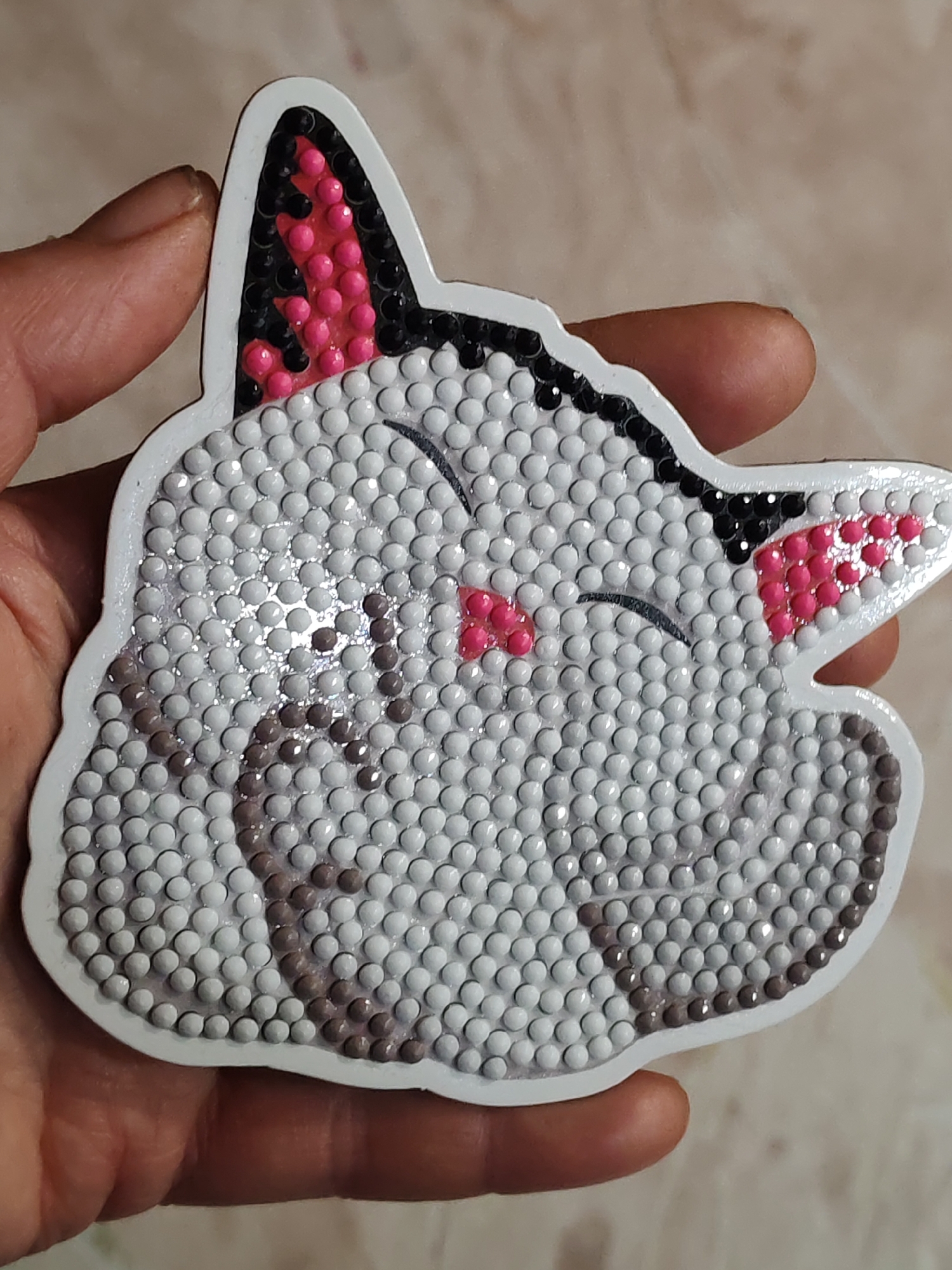 A close-up shot shows a hand holding a cat-shaped object adorned with small, round beads. The cat has white beads covering the majority of its face, with grey beads forming a tabby pattern. The ears are filled with pink and black beads. The nose is also pink, and black beads form the outline of closed eyes. The entire cat shape has a white border, and the background is a light beige. No location in the city of Unknown, Unknown, is visible.