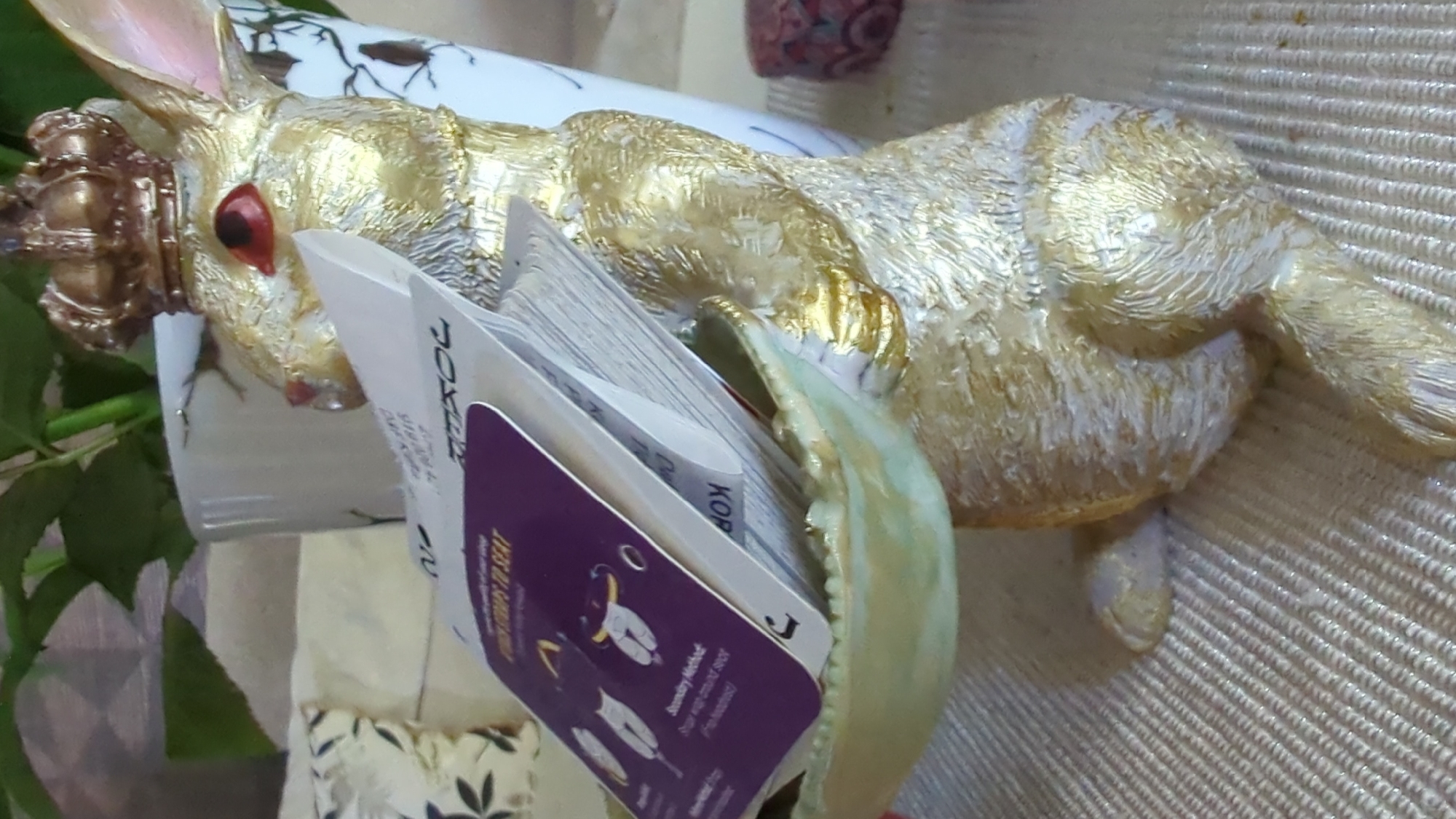 A close-up shot focuses on a golden, textured rabbit figurine wearing a golden crown and holding a light green, bowl-like object in its paws. The figurine has red eyes and is positioned in front of a white vase with a subtle black floral pattern. Several pieces of paper or cards are tucked into the bowl. In the background, hints of greenery, possibly from a plant, are visible. The setting appears to be indoors, with a textured, light-colored surface beneath the figurine. The image is of a decorative object, possibly a candy dish or an art piece. The location is not specified in the image.