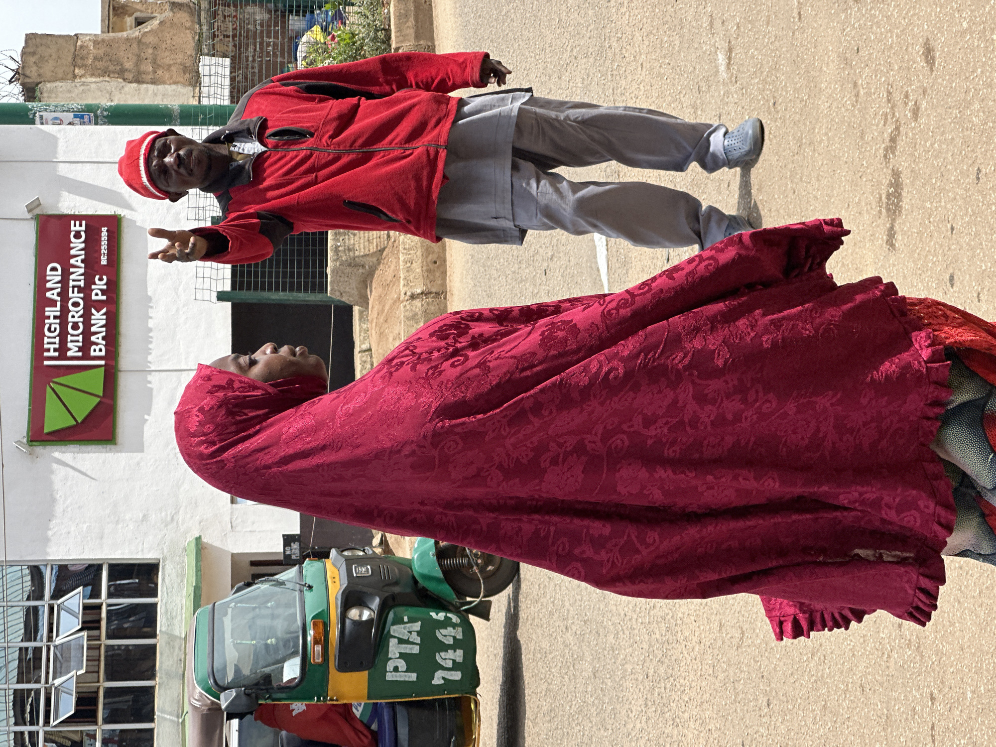 A medium shot captures a street scene in Bukuru, Nigeria, where a woman in a vibrant red hijab walks in the foreground, her face obscured by the garment. Her voluminous hijab features a raised, textured pattern and ruffled edges. Behind her, a man in a red jacket and grey trousers stands gesturing with his right hand, his fingers extended in what appears to be a "peace" sign. He wears a red cap and dark sunglasses. A green and yellow auto-rickshaw, with "PTA 7445" written on its side, is parked to the left. The background shows the white facade of Highland Microfinance Bank Plc, with its green and red logo. The ground is paved with asphalt. The overall lighting suggests it is daytime.