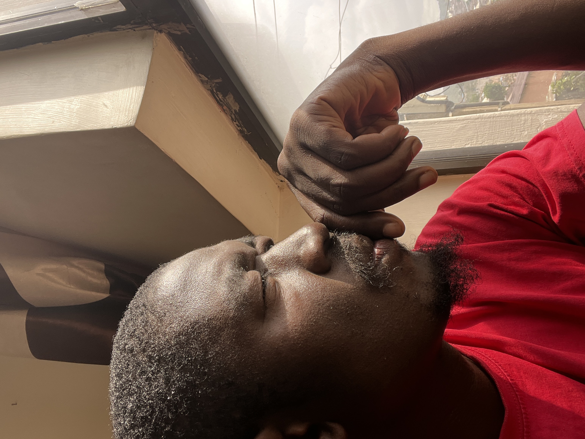 The image is a close-up, profile view of a Black man wearing a red t-shirt, looking out a window. His left hand is raised to his face, with his fingers resting on his chin and mouth. The man has short, dark, curly hair and a light beard. Sunlight streams in from the right, illuminating his face and casting a shadow across the wall. The background shows a window frame and a blurred view of a city with buildings and greenery, likely in Jos, Nigeria.