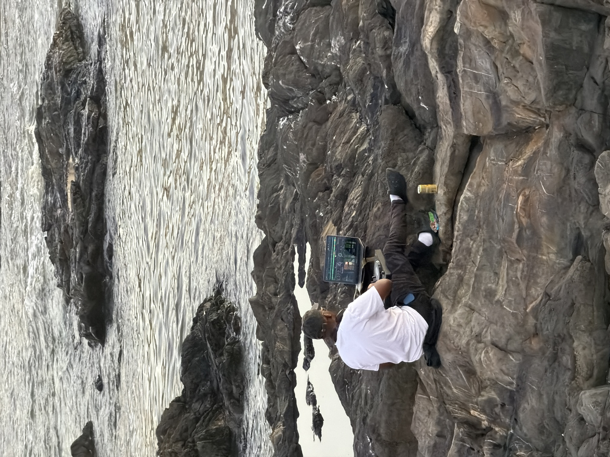 A man in a white shirt and dark pants is rock climbing on a grey rock face. He has a laptop attached to his side and is wearing dark socks and shoes. A half-empty beer bottle and some climbing gear are visible nearby on the rock face. The rock face is located next to a body of water. Based on the style of rock and ocean views, the location is likely along the coastline in Cape Town, South Africa.