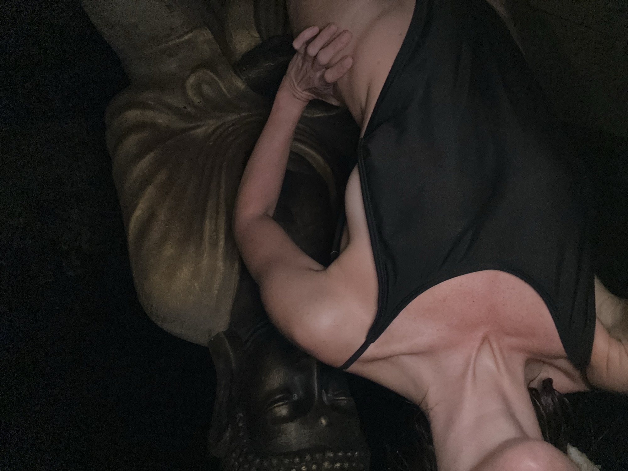 A woman in a black swimsuit is posed next to a dark-faced Buddha statue, with her hand on the statue. The setting is dimly lit and appears to be indoors or in a sheltered outdoor area, possibly in a private residence or a temple setting in Ubud, Indonesia. The woman's skin is visible on her neck, shoulders, and torso. The statue is made of a dark material, possibly stone or resin, with gold-colored drapery. The overall mood is intimate and mysterious due to the low lighting and the juxtaposition of the two figures.