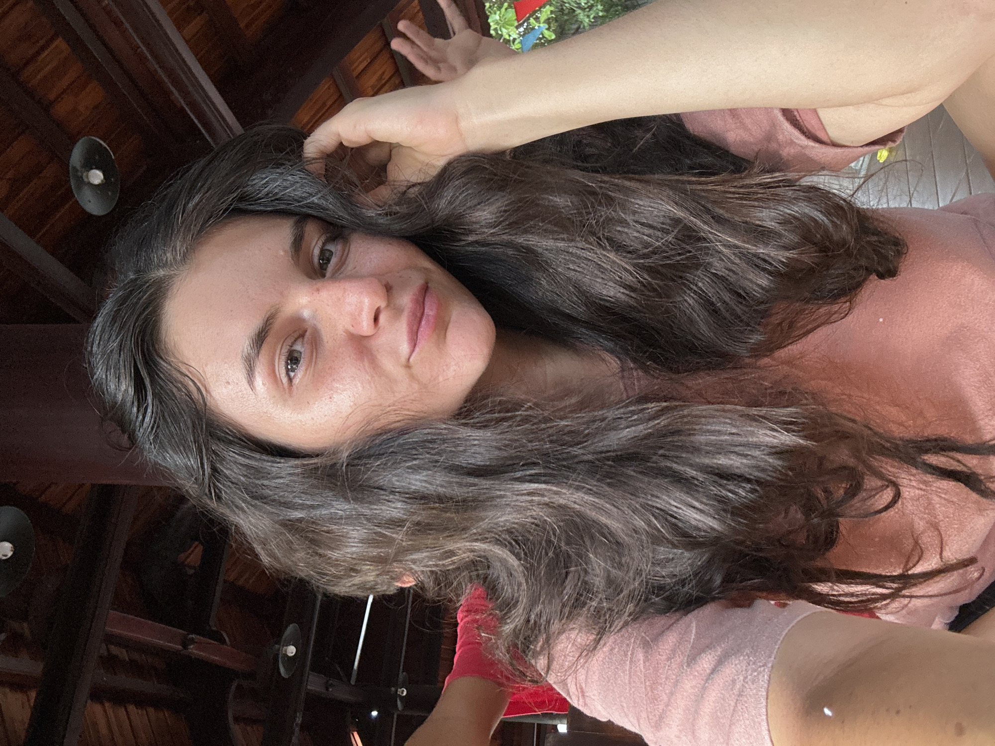 A young woman with long, dark, wavy hair is captured in a close-up, selfie-style portrait, her face turned slightly towards the camera. She is wearing a pink t-shirt. Her right hand is raised, resting on her head as she tucks a strand of hair behind her ear. Her expression is relaxed, with a faint smile. In the background, wooden beams and dark ceiling structures are visible, suggesting an outdoor or semi-outdoor setting, possibly a covered patio or balcony. The environment appears to be warm, with hints of greenery visible to the right. The lighting suggests it is daytime. The presence of a ceiling structure with visible beams and recessed lighting indicates an architectural element common in residential or hospitality areas. Given the context of Mũi Né, Vietnam, this setting could be a resort, cafe, or residential property with traditional Vietnamese architectural influences.