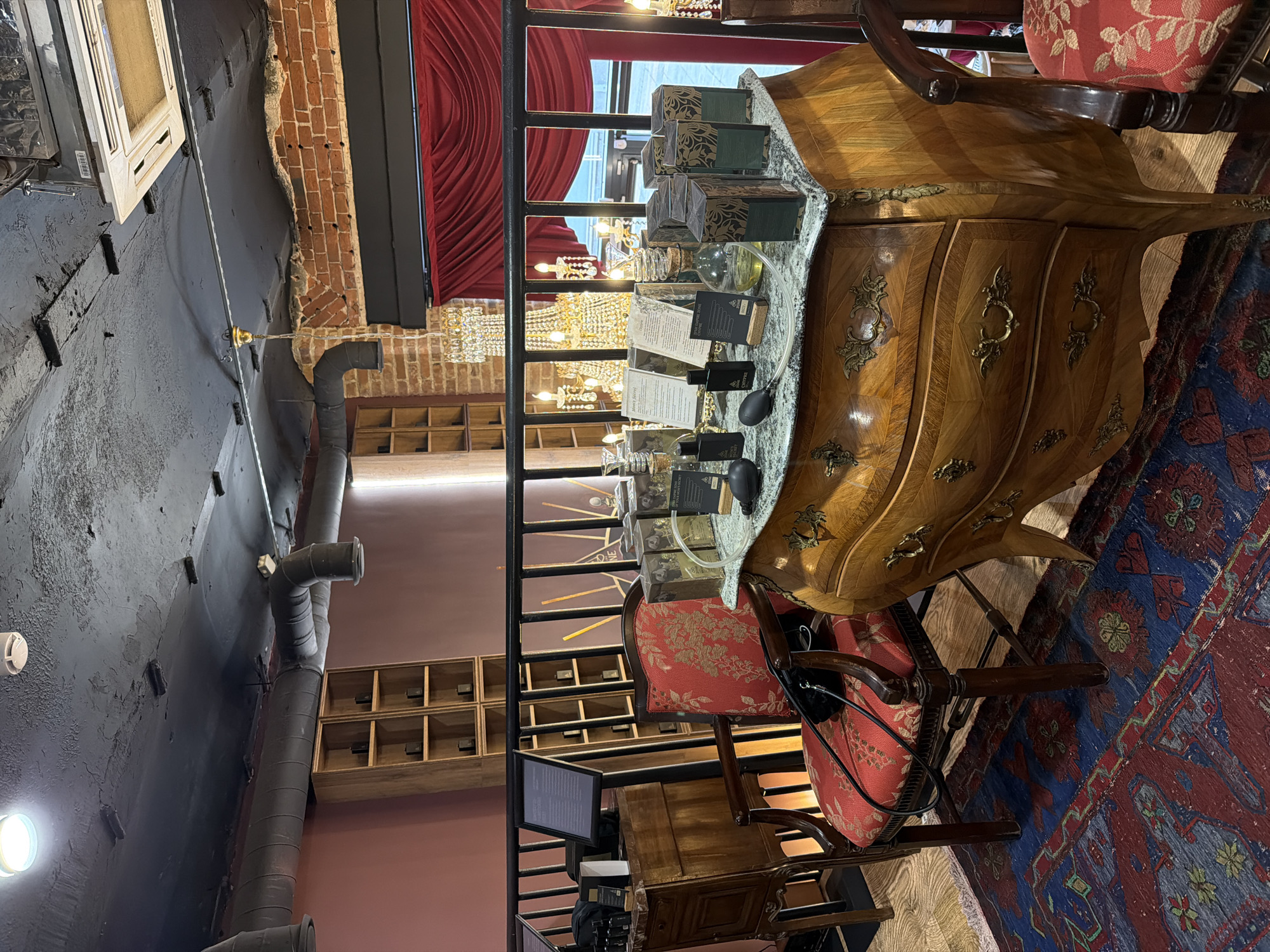 The image depicts an interior space, possibly a shop or a boutique, in what may be Moscow, Russia, given the architectural and decorative elements.
The foreground features an ornate wooden chest or dresser with a curved front and decorative metal hardware. On top of the chest, there are several boxes and bottles, appearing to be perfumes or cosmetics, suggesting the shop's focus.
Next to the chest, there's a wooden chair with a red patterned cushion. Further back, a wrought iron railing partially obscures the view of an area with multiple shelves, some of which contain small, dark objects.
Behind the railing, crystal chandeliers are visible, adding to the luxurious atmosphere. A red curtain partially frames the scene.
The ceiling has exposed ductwork and the walls are a mix of exposed brick and painted surfaces. The flooring is covered by a patterned rug in shades of red, blue, and green.
The image composition is slightly angled, providing an unconventional view of the space. No people are visible in the frame.