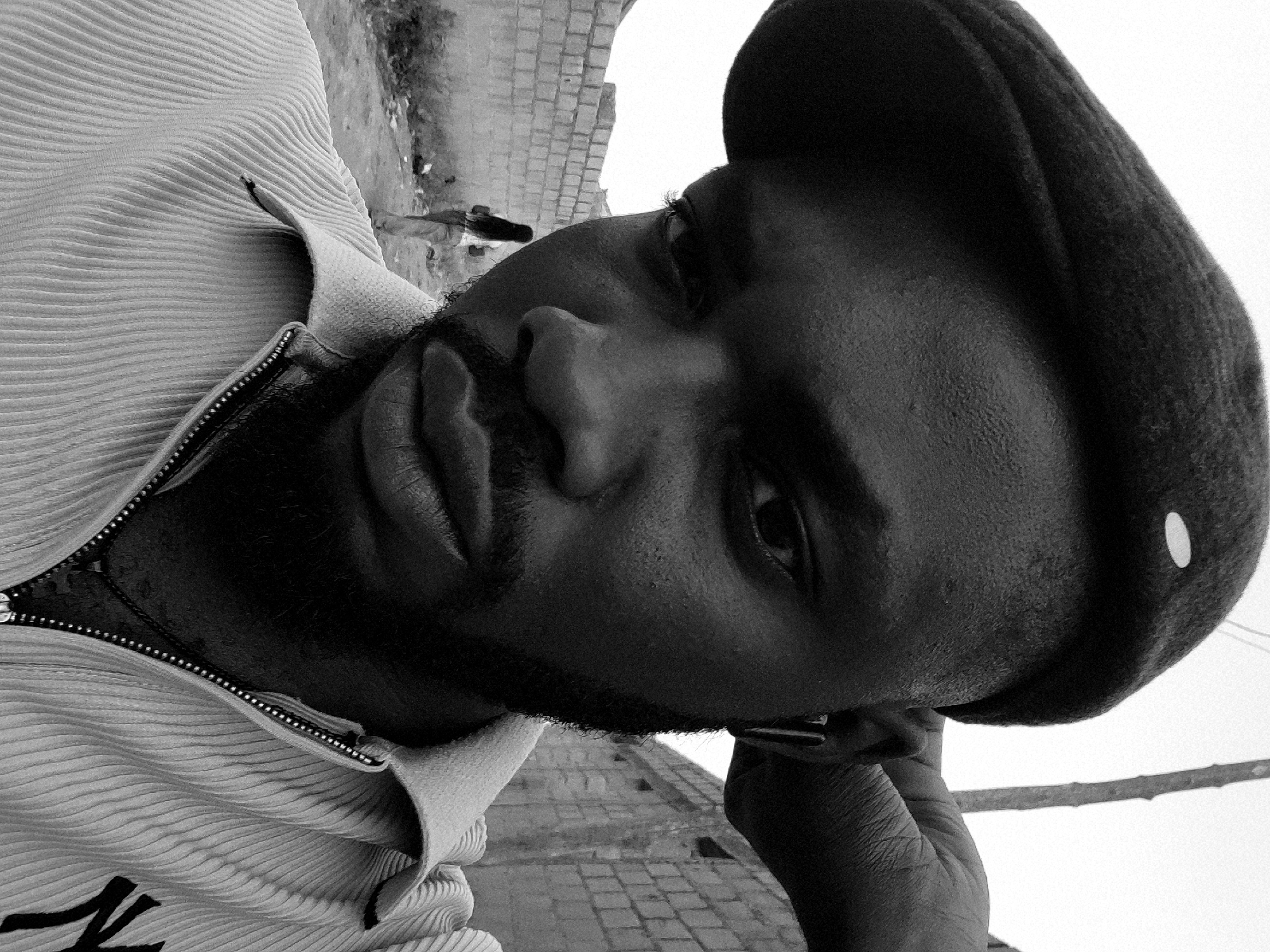The image is a monochrome, close-up portrait of a dark-skinned man wearing a beret and a vertically striped shirt. The man's head is tilted, and he is looking off to the side. His right hand is near his ear. The background includes a brick or stone structure. The specific location is not discernable from the image.