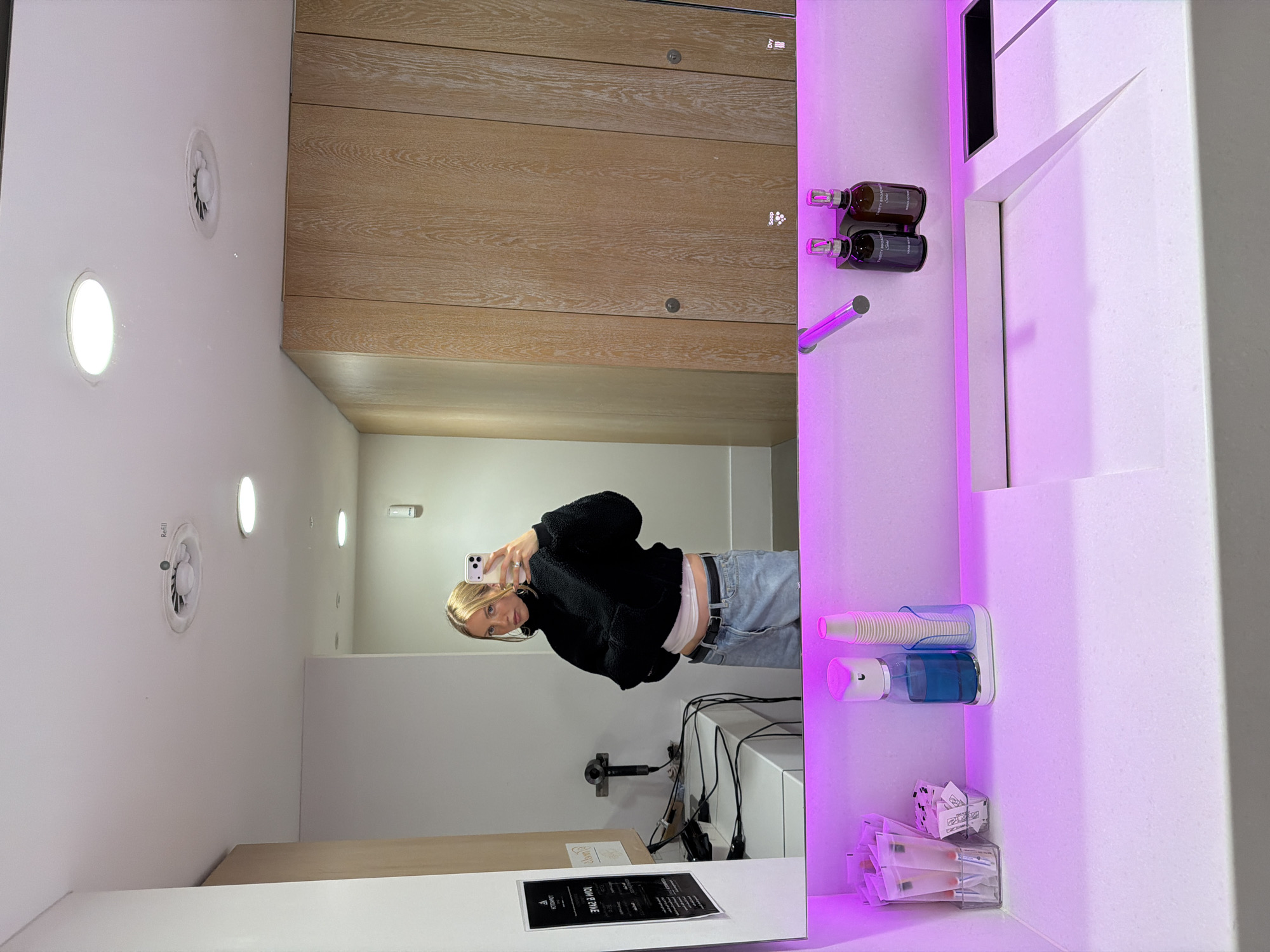 A person is reflected in a mirror, taking a selfie in what appears to be a commercial or public restroom. The individual is wearing a black fleece jacket, jeans, and a white top peeking from under the jacket. The restroom features modern design elements, including wood-paneled walls, recessed lighting, and a vanity area with soap and lotion dispensers. Other items present include hair styling tools, disposable cups, and cosmetic applicators, suggesting the location is possibly in a high-end salon, spa, or upscale retail establishment. The interior design and amenities point towards a location likely found in a more affluent area of London, United Kingdom, though without further context, the precise location is not identifiable.