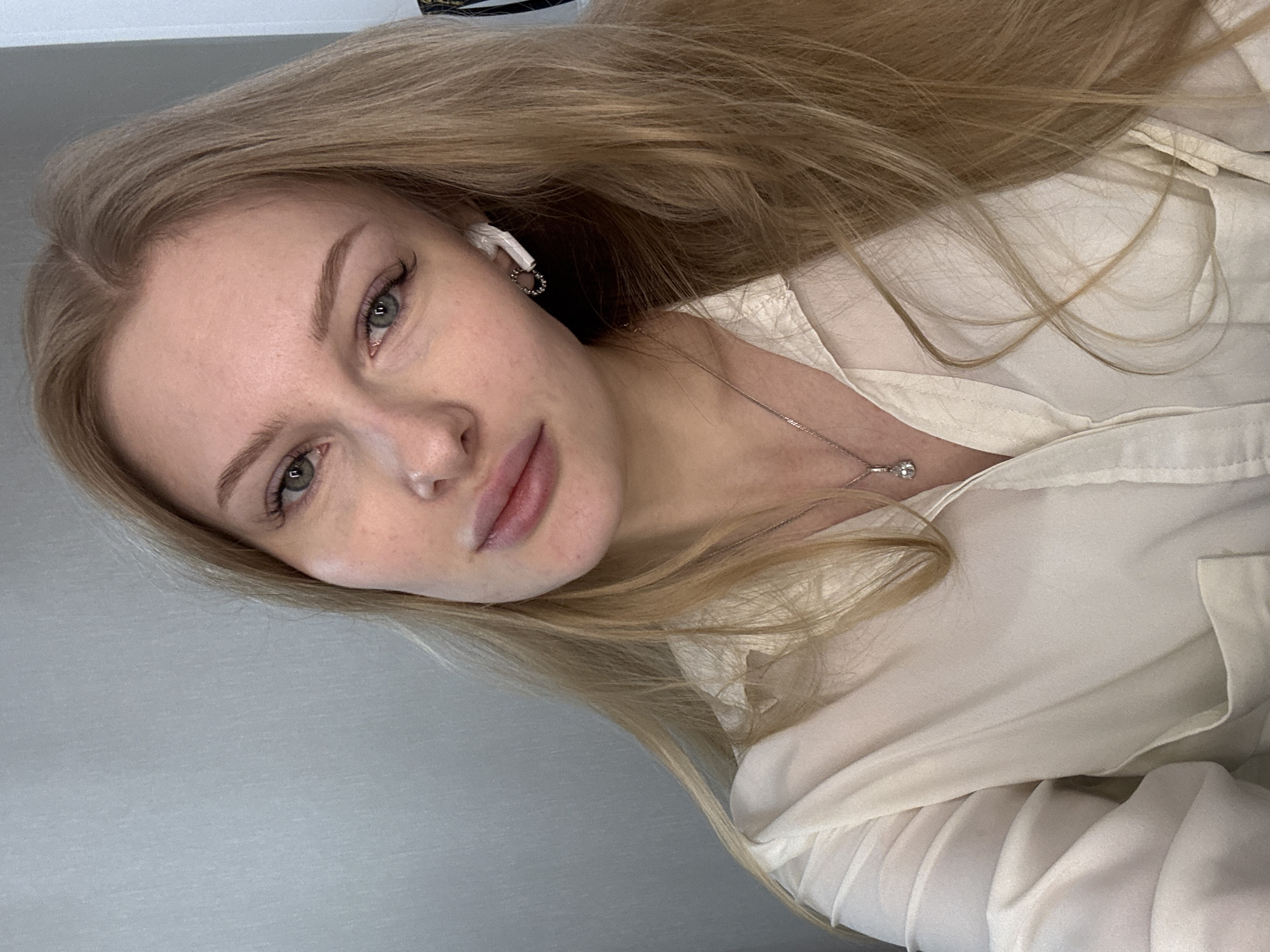 A fair-skinned woman with long, blonde hair is centered in the image. She is wearing a white button-up shirt and a silver necklace with a small pendant. An earbud is visible in her left ear. The background is a plain gray wall. The location is not identifiable as Perm, Russia.