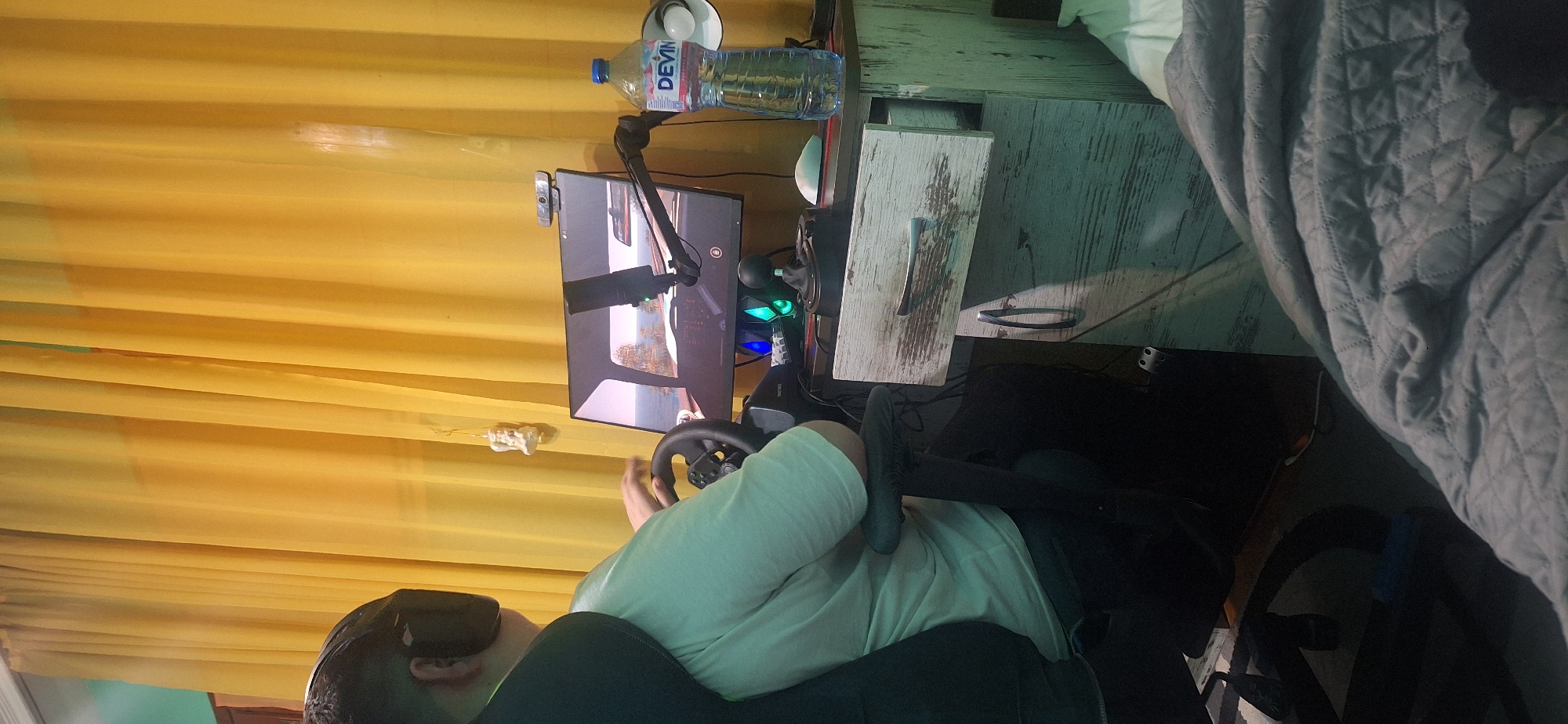 A person wearing a VR headset sits in a gaming chair and uses a steering wheel. The person wears a light green t-shirt. A monitor displays a car racing game, with a microphone attached to the monitor. A water bottle sits on the desk next to the monitor. The room has yellow curtains and a textured gray blanket on a bed. The exact location within Stara Zagora, Bulgaria is indeterminable.