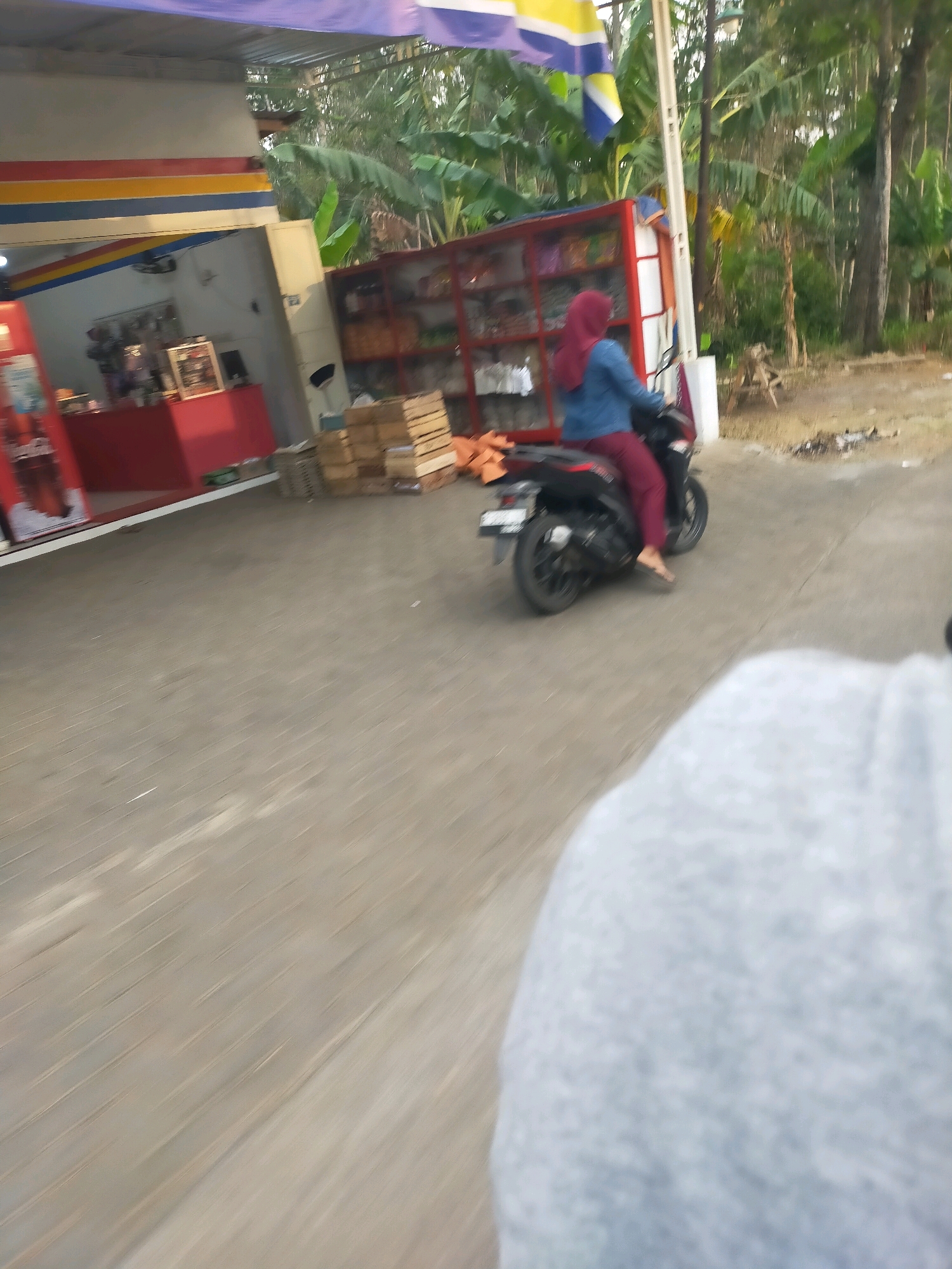 The image captures a scene on a paved road in Harjokuncaran, Indonesia, with a small store visible on the left. A person wearing a maroon hijab and maroon pants is riding a black motorcycle. The motorcycle is in motion, suggested by the blurriness of the wheels. Behind the motorcycle, there's a red display shelf filled with items, and stacks of wooden crates are piled nearby. The environment appears to be semi-rural, with trees and vegetation in the background. The overall lighting suggests it is daytime. The perspective is from another moving vehicle.