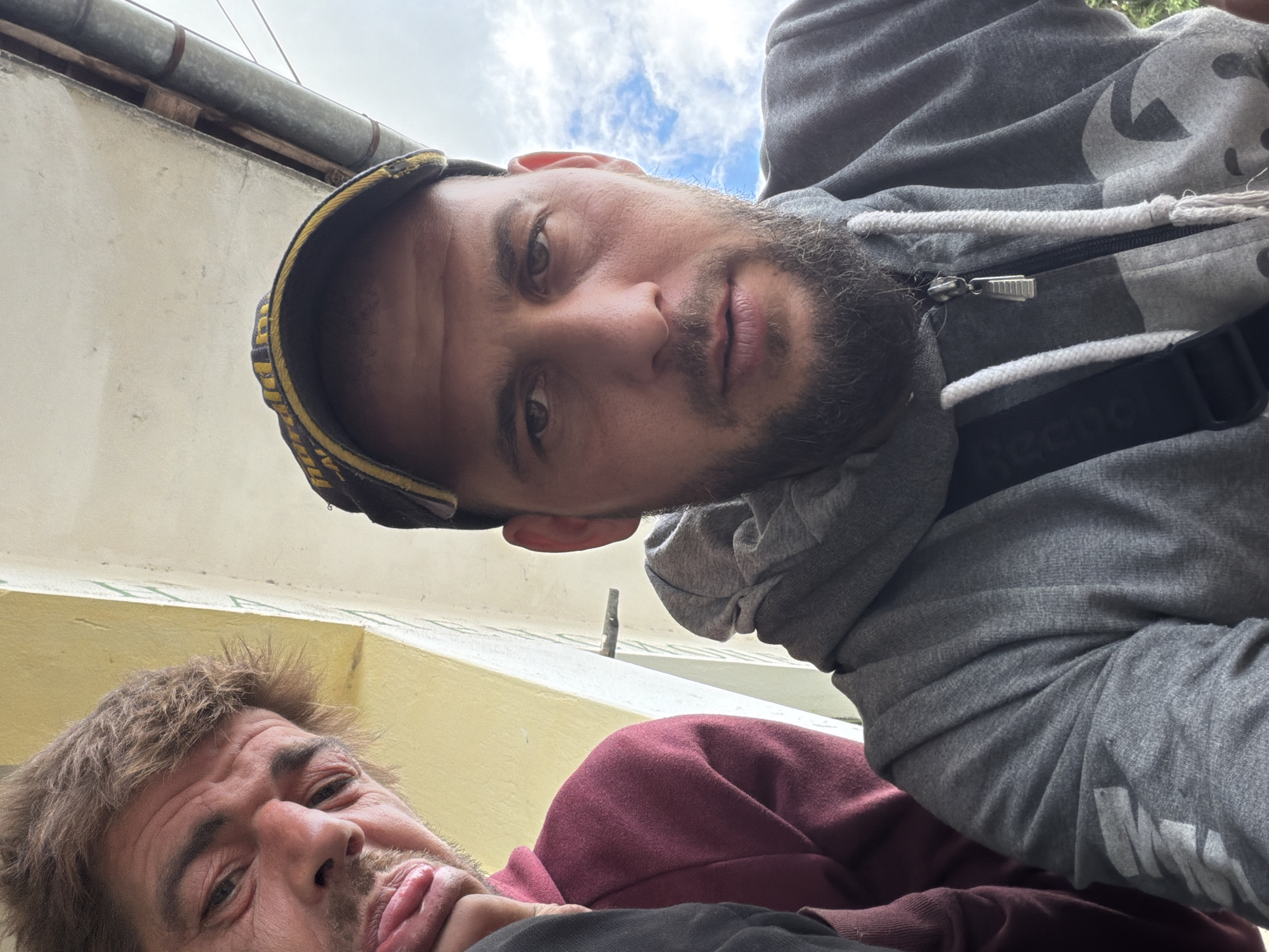 The image is a close-up, low-angle shot featuring two men, suggesting a selfie taken outdoors. The perspective is tilted, looking upwards towards the subjects and the sky.
The primary subject, positioned in the upper part of the frame, is a man with light skin, short dark hair, and a short beard or stubble. He wears a grey hooded sweatshirt with a white drawstring, and a dark cap with yellow or gold detailing on the brim. He looks directly at the camera with a neutral or slightly serious expression. A black strap, possibly from a bag, crosses his chest, with the brand name "Reebok" visible on it.
Below and to the left of the first man, partially obscured, is a second man. He has light skin, messy brown hair, and some facial stubble. He wears a dark red or maroon sweatshirt and is making a humorous or playful face, sticking his tongue out.
The background features a light-colored (possibly beige or light yellow) wall. Above the wall, a metal gutter or drainpipe runs horizontally, with some wooden planks visible behind it. Patches of blue sky with white clouds are visible in the upper background, indicating daylight. On the grey clothing of the first man, towards the bottom right, the word "NIKE" is partially visible. The scene has a casual and informal atmosphere. Given the context of Sirakovo, Bulgaria, it suggests a candid moment in an everyday outdoor setting.