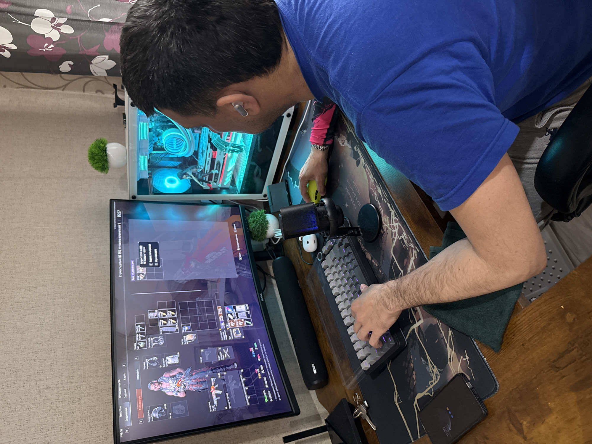 A man is playing a computer game. He is wearing a blue shirt and gray sweatpants. He is sitting at a wooden desk with a computer, monitor, keyboard, mouse, and microphone. The monitor displays a game interface showing inventory and character. His computer tower, featuring teal-lit internal components, sits next to the monitor, alongside a small potted artificial plant. A dark mousepad and a mobile device are also visible on the desk. The background shows a curtain with a flower design. The OCR indicates the game display details and mentions the option to enable or disable character display during raids, suggesting this is a gaming setup within a residential environment. No specific details pinpoint the location to Rusanovo, Russia.