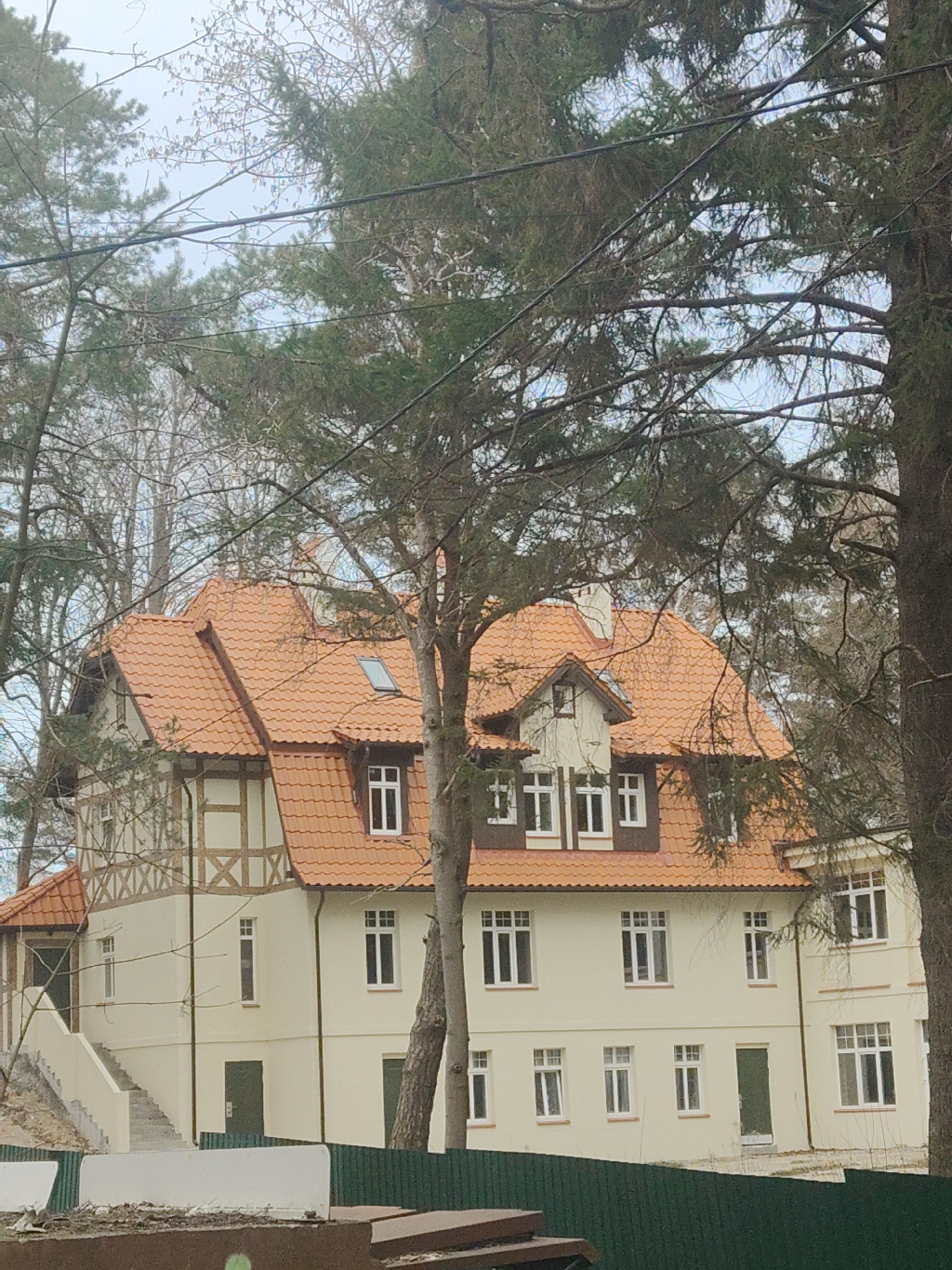 The image shows a three-story building with a red tile roof. The building is painted in a light beige color and has white-framed windows. There are trees in the foreground, partially obscuring the building. The location is Unknown, Unknown. A green fence runs along the bottom of the frame. Construction materials are visible in front of the fence.