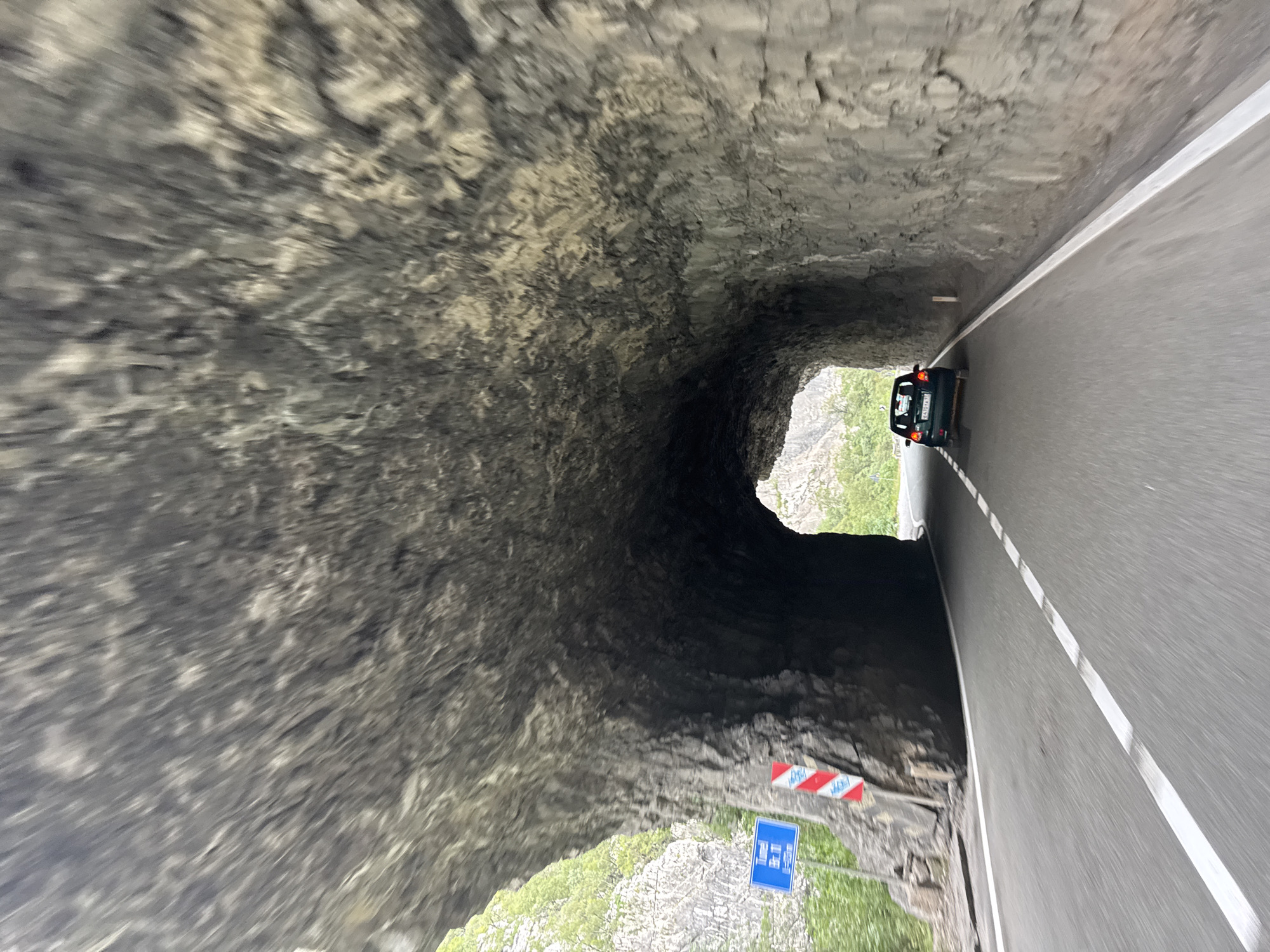 The image shows a dark, rocky tunnel carved into a mountainside. A dark-colored car is driving through the tunnel on a paved road marked with white lines. The car is in the right lane of the two-lane road. The tunnel ceiling is rough and textured, with visible rock formations. Outside the tunnel entrance, on the left side of the road, there is a blue sign with white text and a red and white striped marker indicating a hazard. Green vegetation is visible on the mountainside outside the tunnel. The image is slightly blurred due to motion, suggesting it was taken from a moving vehicle. The scene is consistent with the mountainous terrain found near Vlahovići, Montenegro, known for its tunnels and natural beauty.