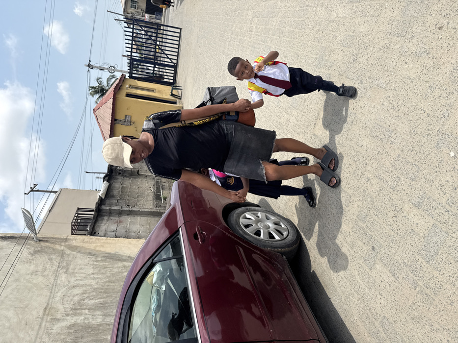 This image captures an outdoor scene in Ado, Nigeria, featuring an adult female walking with two children. A maroon sedan is parked on the left, its rear passenger side visible. The adult female, positioned centrally, wears a beige baseball cap, black short-sleeved top, dark denim skirt, and grey slide sandals. She carries a dark shoulder bag with yellow accents and a grey cooler bag. She holds the left hand of a young female child, partially obscured behind her, who wears a dark school uniform. Simultaneously, she holds the right hand of a young male child on her right, who is dressed in a white short-sleeved school shirt, a red tie, dark trousers, dark athletic shoes, and a yellow backpack. The male child looks towards the camera with a slight smile as he walks.
The background features an unpainted concrete building with a satellite dish attached to its upper exterior on the far left. A concrete perimeter wall separates this building from a small, yellow-painted security post with a red corrugated roof, positioned further to the right. Beyond the security post, a large, dark metal gate, appearing partially open, leads to another area. Utility poles and overhead power lines are visible against a bright blue sky with scattered white clouds. The ground is a light-colored, paved surface, and distinct shadows are cast from the subjects, indicating strong overhead sunlight.