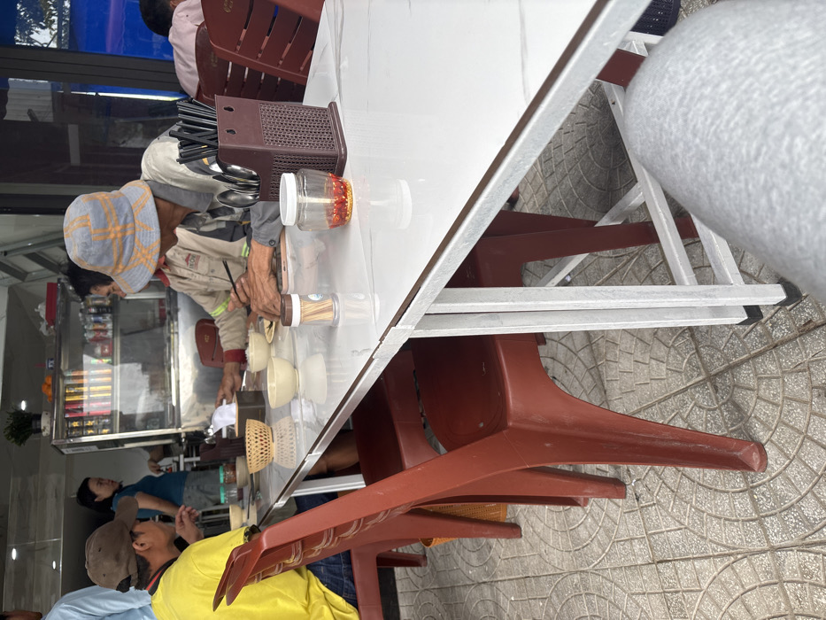 The image depicts a casual open-front eatery in Ngũ Hành Sơn Ward, Vietnam, during daylight. A white rectangular table with a light grey marble-patterned surface occupies the foreground and mid-ground, supported by white metal legs. Four individuals are visible, with the perspective taken from a fifth person whose leg, clad in grey fabric, is seen in the lower right.
On the right side of the table, a male subject wearing a grey and orange checkered bucket hat and a light-colored long-sleeved shirt is seated, actively using black chopsticks to eat from a small, light-colored ceramic bowl. A smartphone is near his right hand. Opposite him, on the left, a male subject in a yellow short-sleeved shirt and a brown baseball cap is partially visible, also seated at the table.
Table accessories include a dark brown plastic holder containing black chopsticks and silver spoons, a clear glass jar with red chilies or chili flakes and a white lid, a white toothpick dispenser with a brown lid, a dark brown tissue box cover with a white tissue, and a light brown woven basket. Several empty small, light-colored ceramic bowls are also present. The chairs are dark red/brown plastic. The floor is covered with grey interlocking paving tiles.
In the background, a female subject in a blue t-shirt stands behind a glass display counter, facing the diners. A green potted plant is on top of this counter. A partial view of a person in a light blue shirt is visible on the far left, and another person in a pink shirt is partially seen at a distant table on the far right.