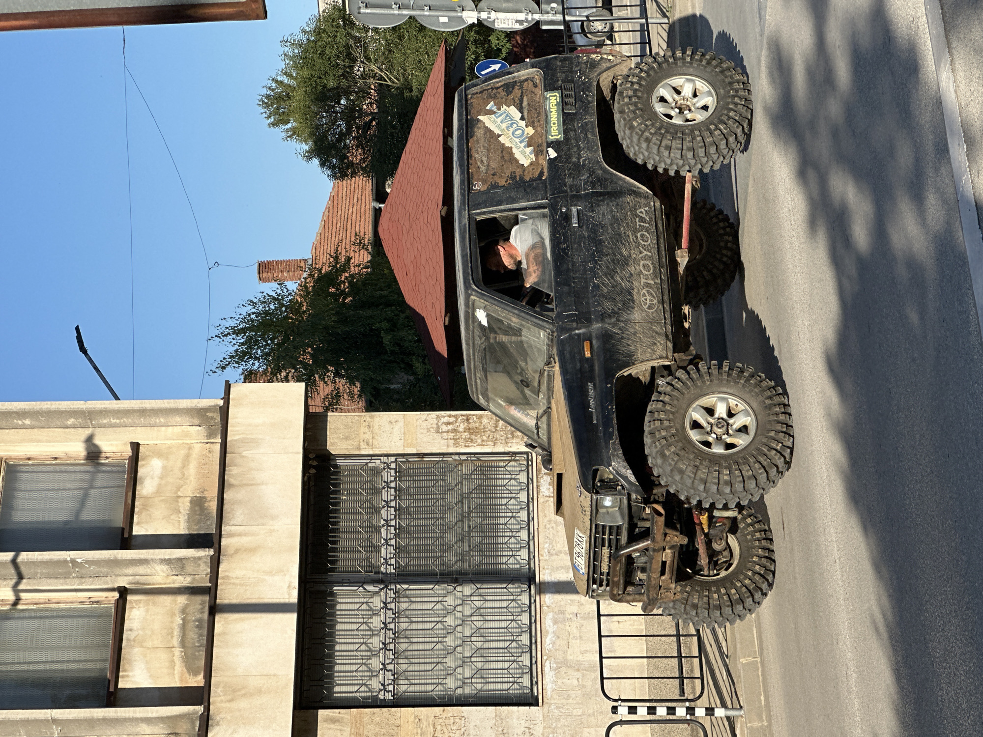 The image captures a modified black Toyota Land Cruiser J80 series, equipped with oversized off-road tires and a reinforced front bumper, parked on a paved street in Lyaskovets, Bulgaria. The vehicle is dirty, suggesting recent off-road use. A middle-aged man with a beard, wearing a grey t-shirt, is visible inside the driver's side, holding the steering wheel. The vehicle has stickers on its rear window, one appearing to be a map of an area with Cyrillic text and another that reads "IRONMAN."
To the left of the vehicle, a building with a beige facade features two large windows with decorative metal grates. The building's architecture suggests it is an older structure, possibly residential or commercial. Sunlight casts shadows from the building and trees onto the street.
In the background, a red-tiled roof of another building is visible, along with green trees and a clear blue sky. Traffic signs and a lamppost are also present. The overall scene depicts a moment in a town street, highlighting the rugged appearance of the modified vehicle against an urban backdrop in Lyaskovets, Bulgaria.