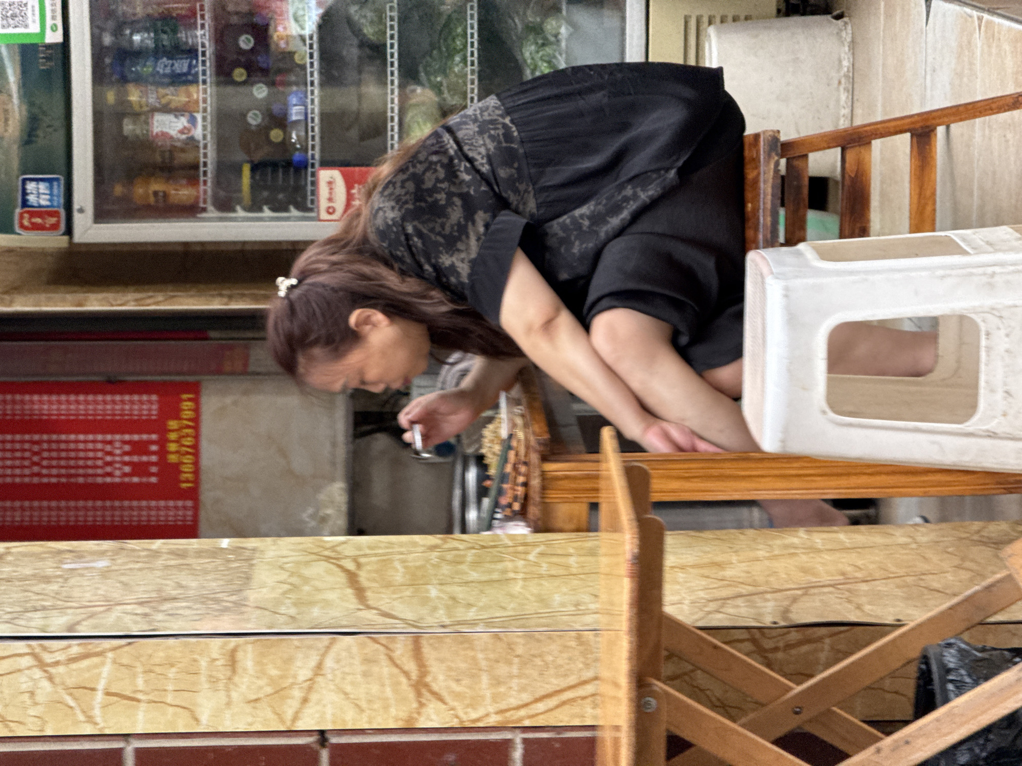 A woman in Jiangjin, China, wearing a dark patterned dress and with her hair pulled back, bends forward while standing behind a tan counter. She holds a metallic object in her right hand. To her left is a tall cooler filled with beverages. Behind her, there is a wooden chair and a white plastic stool. The exterior of the building features a red sign with Chinese characters and a phone number. A wooden folding chair is visible in the foreground. A black garbage bag is on the lower right.