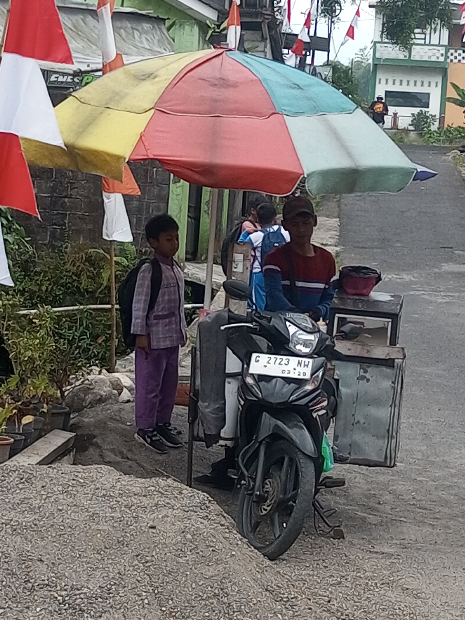 In Krasak, Indonesia, a young boy wearing a purple patterned shirt and pants stands with a black backpack on his left shoulder, facing right. He is positioned under a large, multicolored umbrella. To his right, another boy, wearing a dark baseball cap and a maroon shirt with blue and white stripes, stands behind a modified motorcycle. This motorcycle has a metal box attached to its right side and a license plate reading "G 2723 NW". The background shows a street with buildings, including one with green trim, and trees. Several Indonesian flags, red and white, are visible on poles. A pile of gravel is in the foreground.