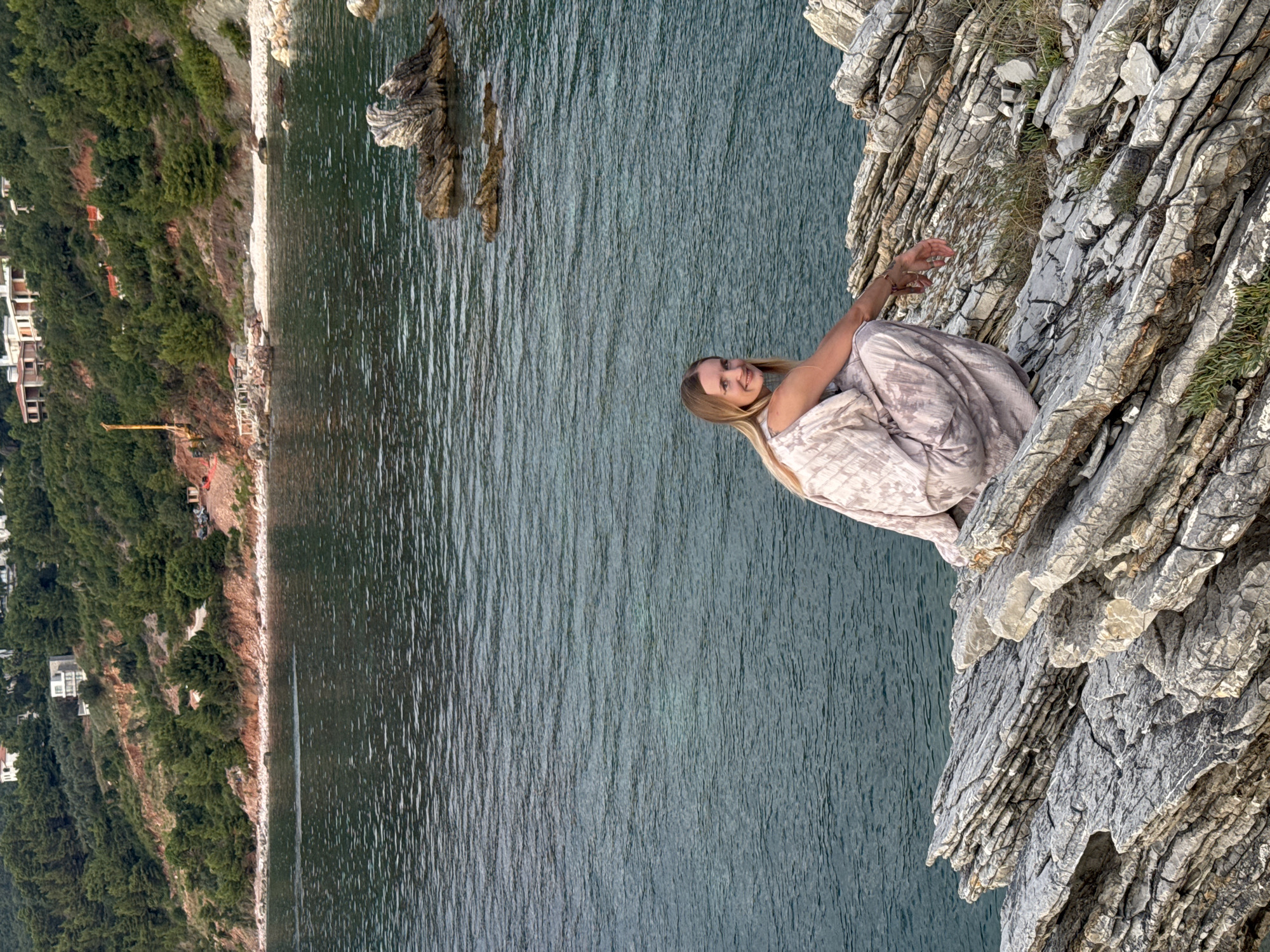 In Sveti Stefan, Montenegro, a blonde woman wearing a light-colored, patterned dress crouches on layered, grey rocks overlooking a body of water. The water is a deep teal color with visible ripples. In the background, a green, tree-covered hillside rises, with several white buildings constructed on its upper slopes. The coastline features a mix of sand and smaller rocks. The sky is not visible in the frame. The overall scene depicts a natural coastal landscape with human settlement in the distance.