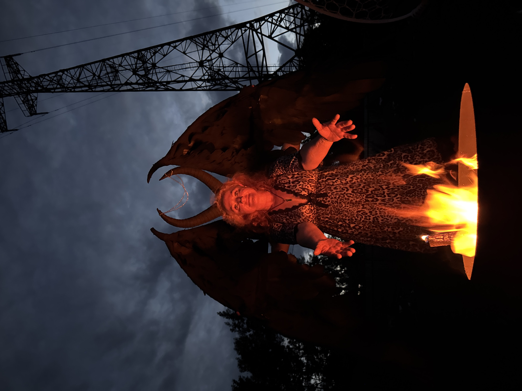 An adult with curly blonde hair, wearing a leopard print dress and a necklace, stands in front of a campfire, holding their arms out. The adult has large, dark, horned wings and horns on their head. A tall, metal power line tower is visible in the background. The sky is dark and cloudy, suggesting it is either dusk or dawn. The location is identified as Chaikovsky, Russia.
