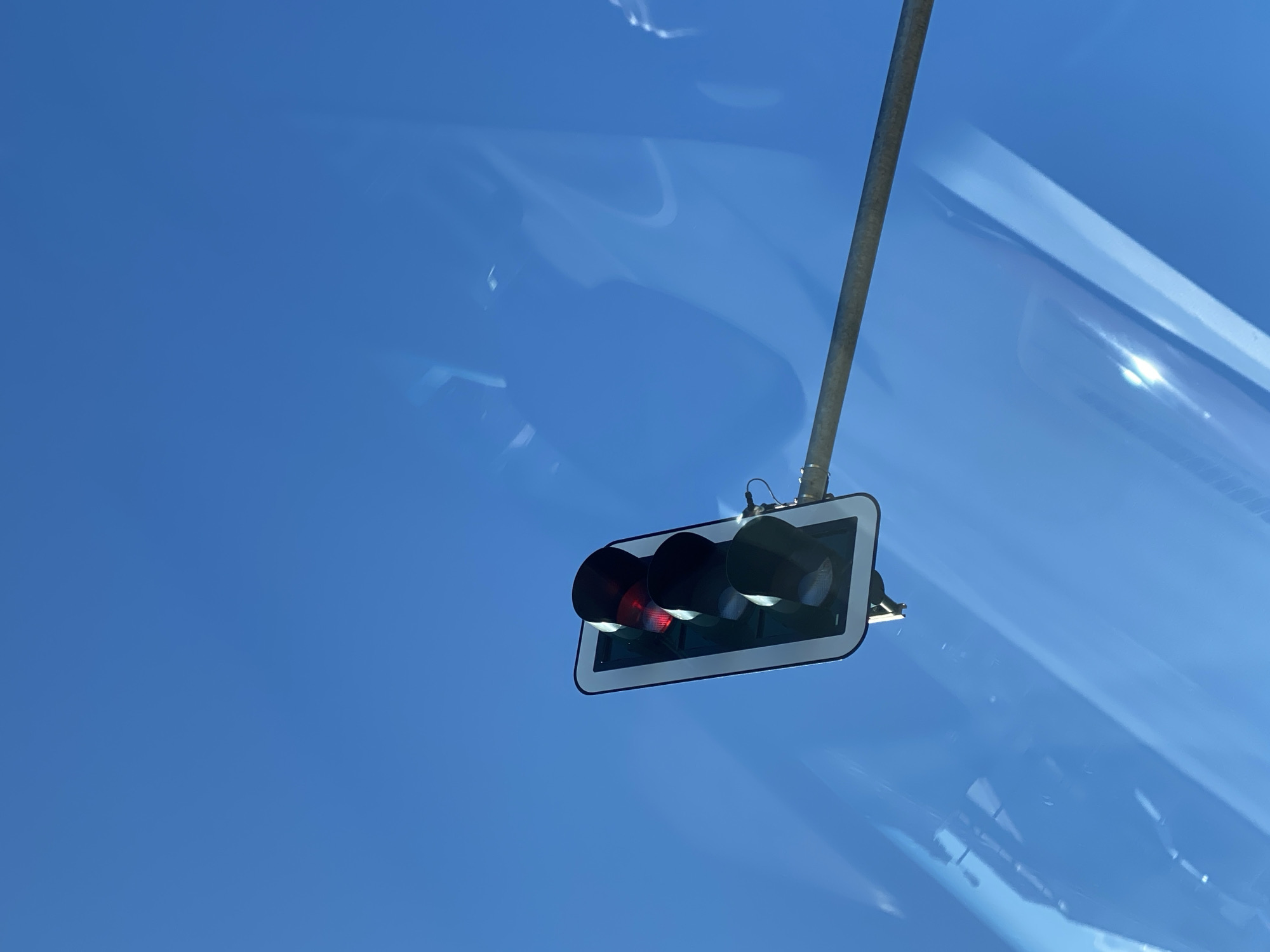 A traffic light with its red light illuminated is mounted on a metal pole against a clear blue sky. The traffic light is positioned to the right of the frame, angled upwards. Reflections and light patterns are visible on the surface of the traffic light's casing and within the sky itself, suggesting the presence of a reflective surface such as glass. The context provided indicates this scene is located in Werther, Germany.