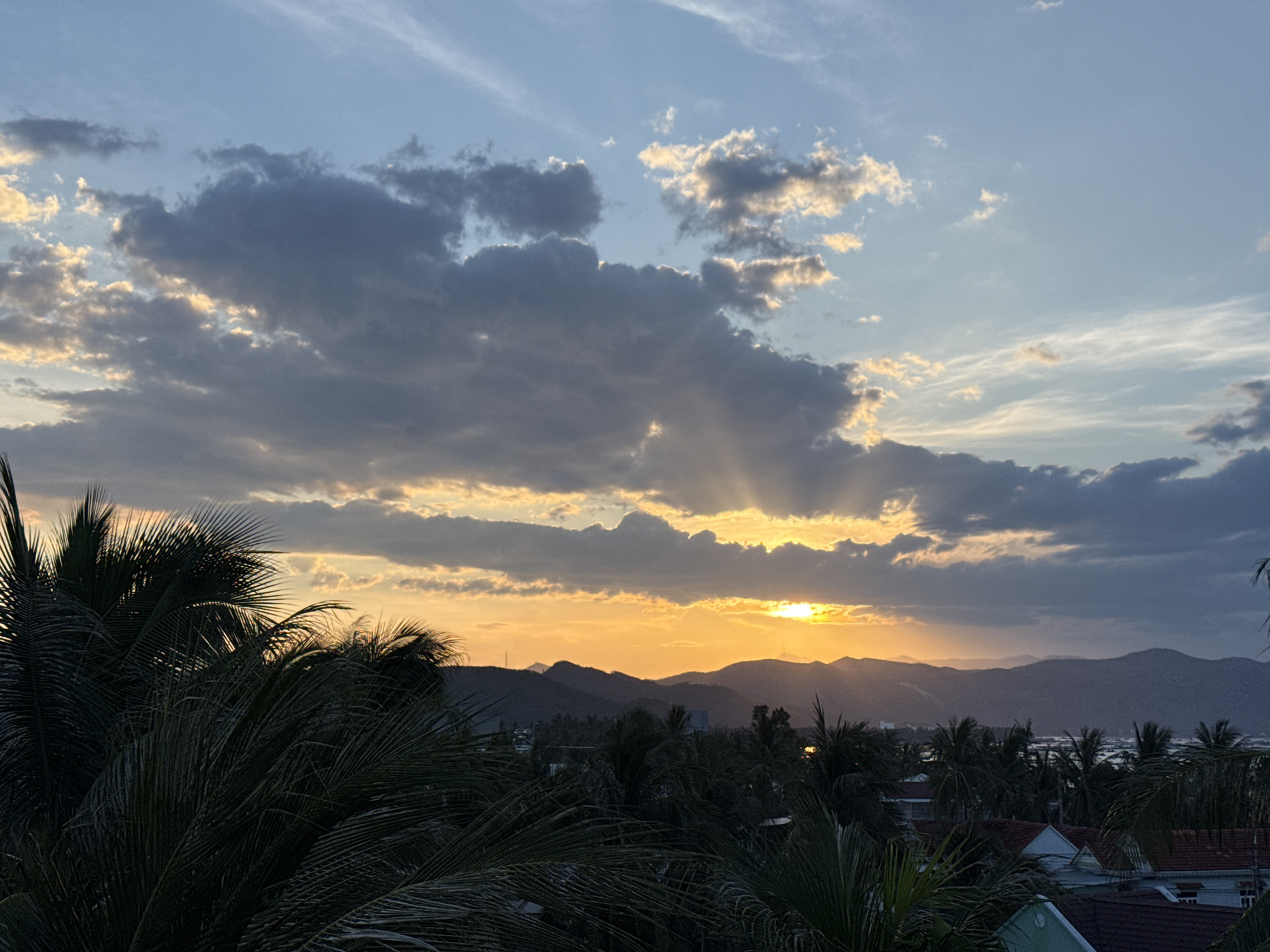 A wide-angle, eye-level shot captures a serene sunset over a tropical landscape. The sky is a canvas of soft blues and grays, with dramatic rays of sunlight piercing through dark, textured clouds. In the foreground, lush, dark green palm fronds frame the scene, their delicate leaves silhouetted against the vibrant sky. Beyond them, a range of dark, layered mountains stretches across the horizon, partially obscuring a distant body of water that reflects the warm hues of the setting sun. A cluster of buildings with reddish-brown tile roofs peeks through the dense foliage of palm trees, suggesting a coastal town or village nestled within this tropical paradise. The overall atmosphere is peaceful and tranquil, evoking the beauty of a tropical evening.