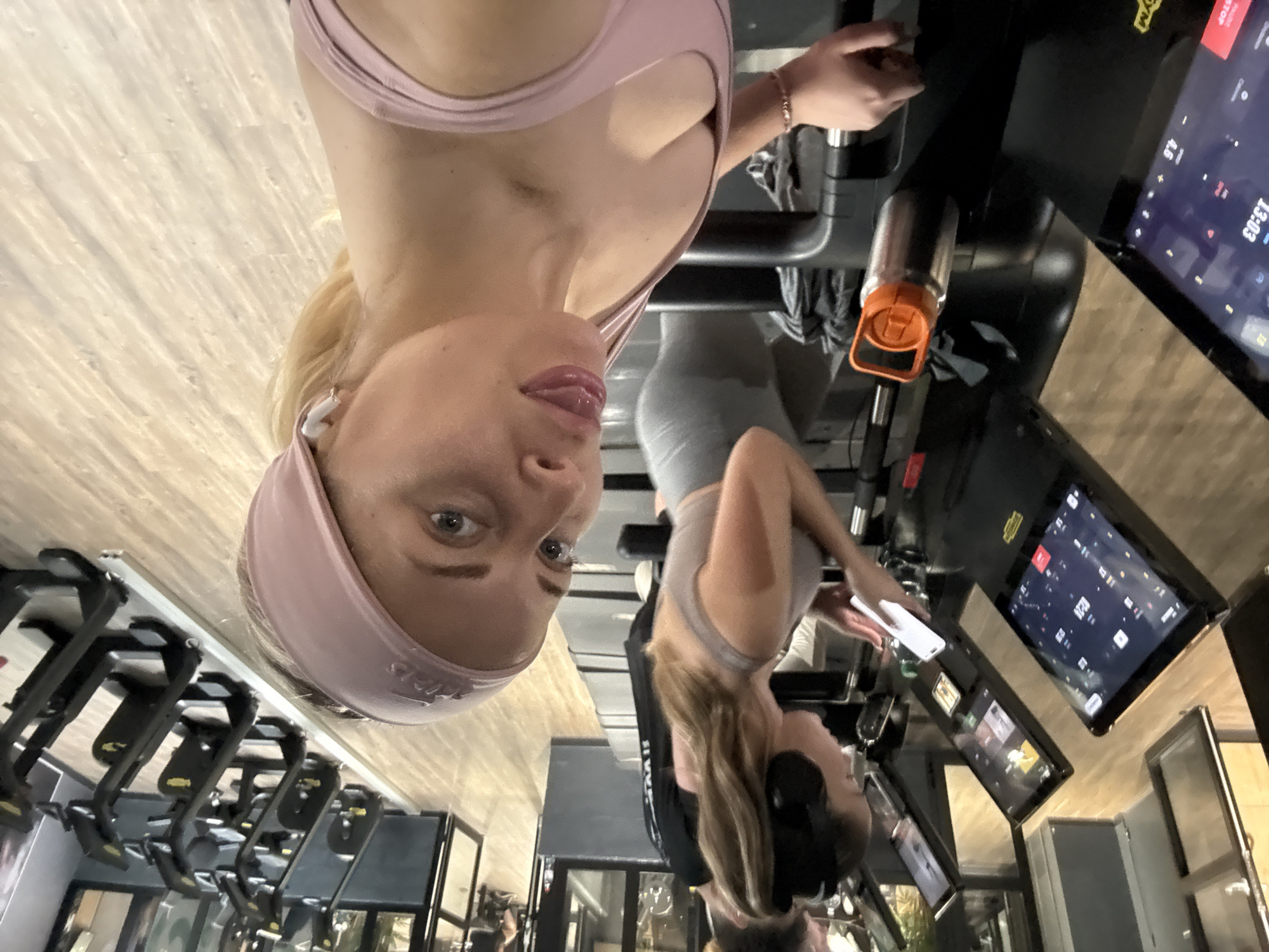 The image is taken inside a gymnasium. A woman in a pink sports bra and headband is positioned in the foreground. Another woman in a grey sports bra and leggings is further away. Gym equipment such as treadmills are visible in the background. The location appears to be Paris, France.