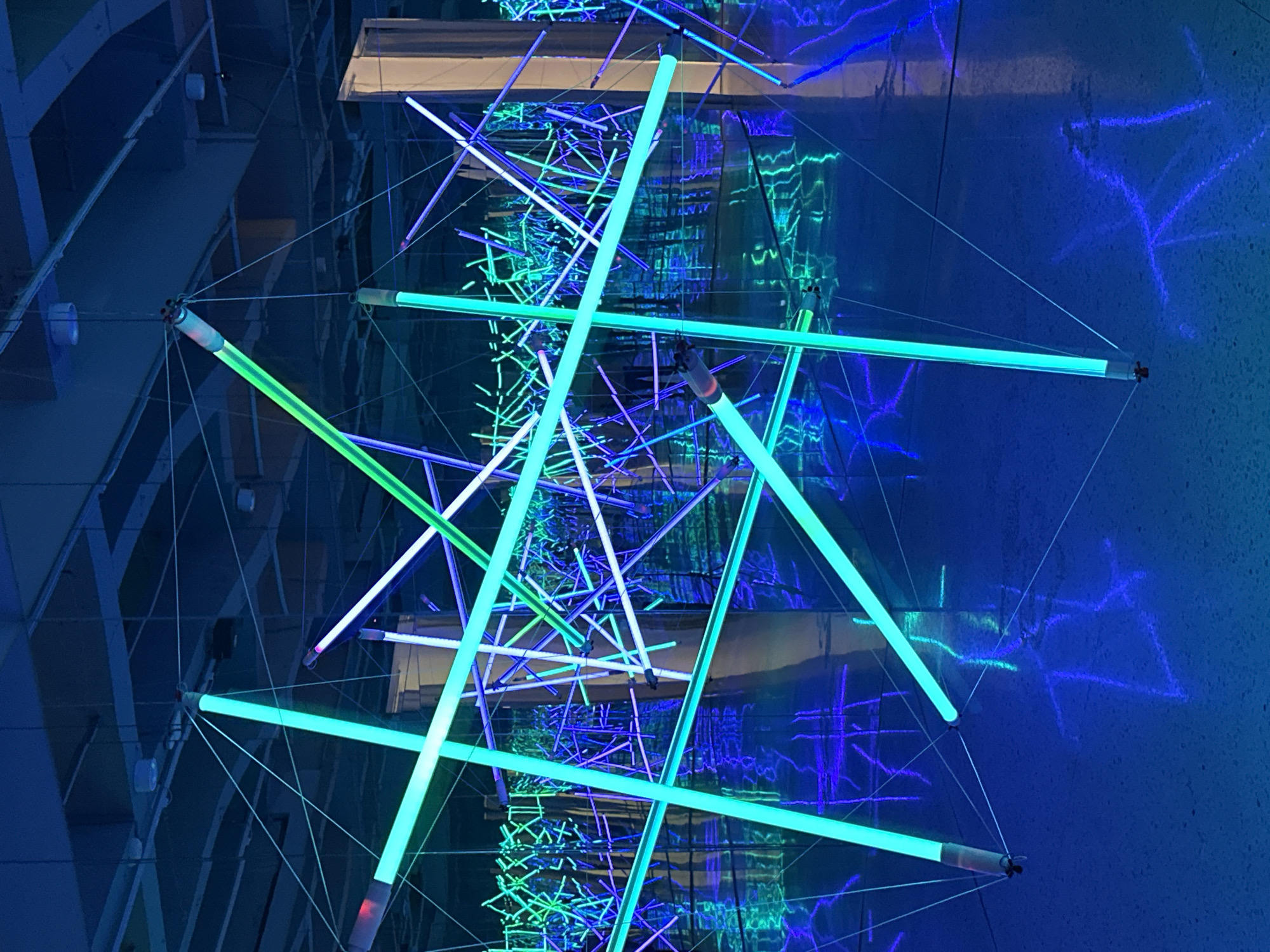 The image shows an art installation with neon lights arranged in geometric patterns, suspended by wires. The predominant colors are shades of blue and green. The background is a dark blue, possibly a wall or reflective surface, with lighter blue designs that appear to be light reflections. The location in the city of Banan, China is unknown from this image alone. There are no people visible in the image.